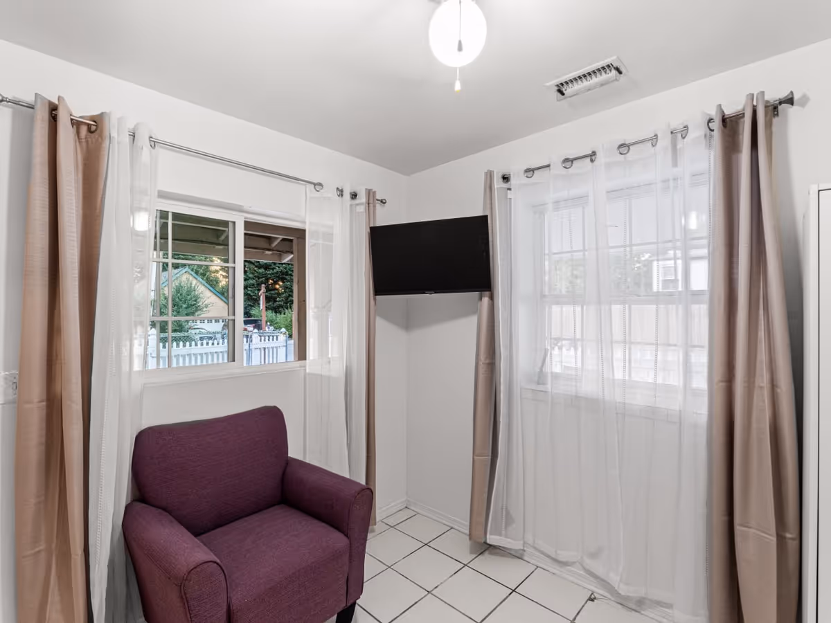 Small living area with purple armchair, two windows with sheer curtains, and a wall-mounted TV.