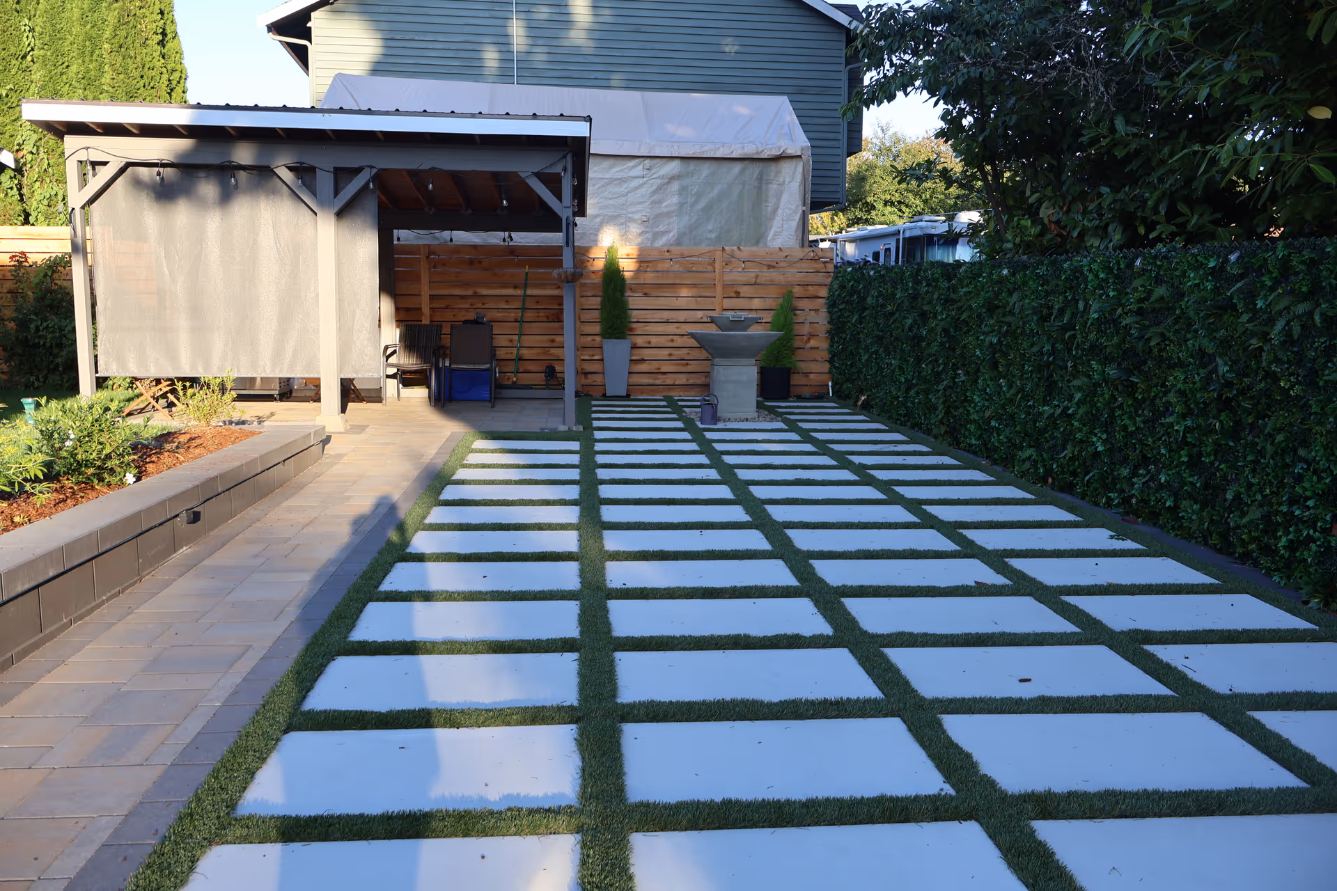 Modern backyard with stepping-stone path, covered seating area, and tall hedges.
