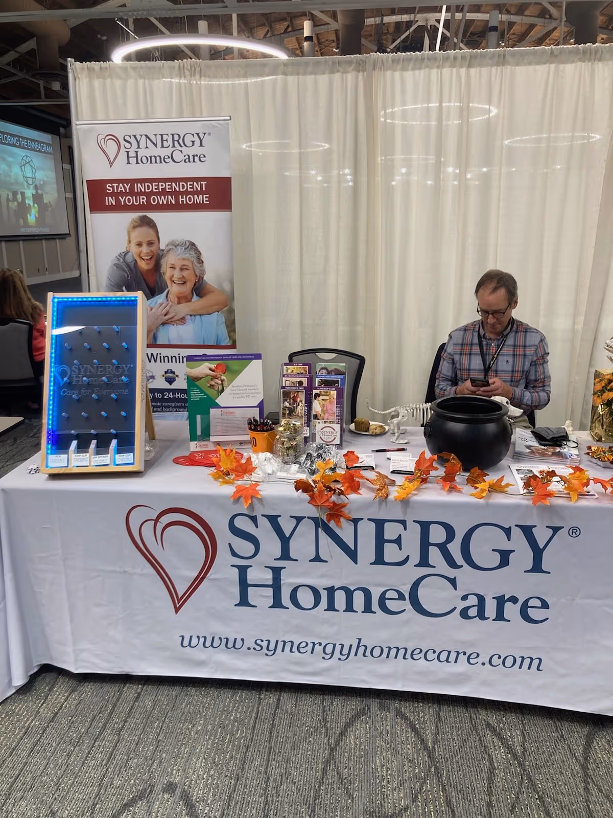 Trade show booth for SYNERGY HomeCare with banner, table, leaves, and a man on his phone.