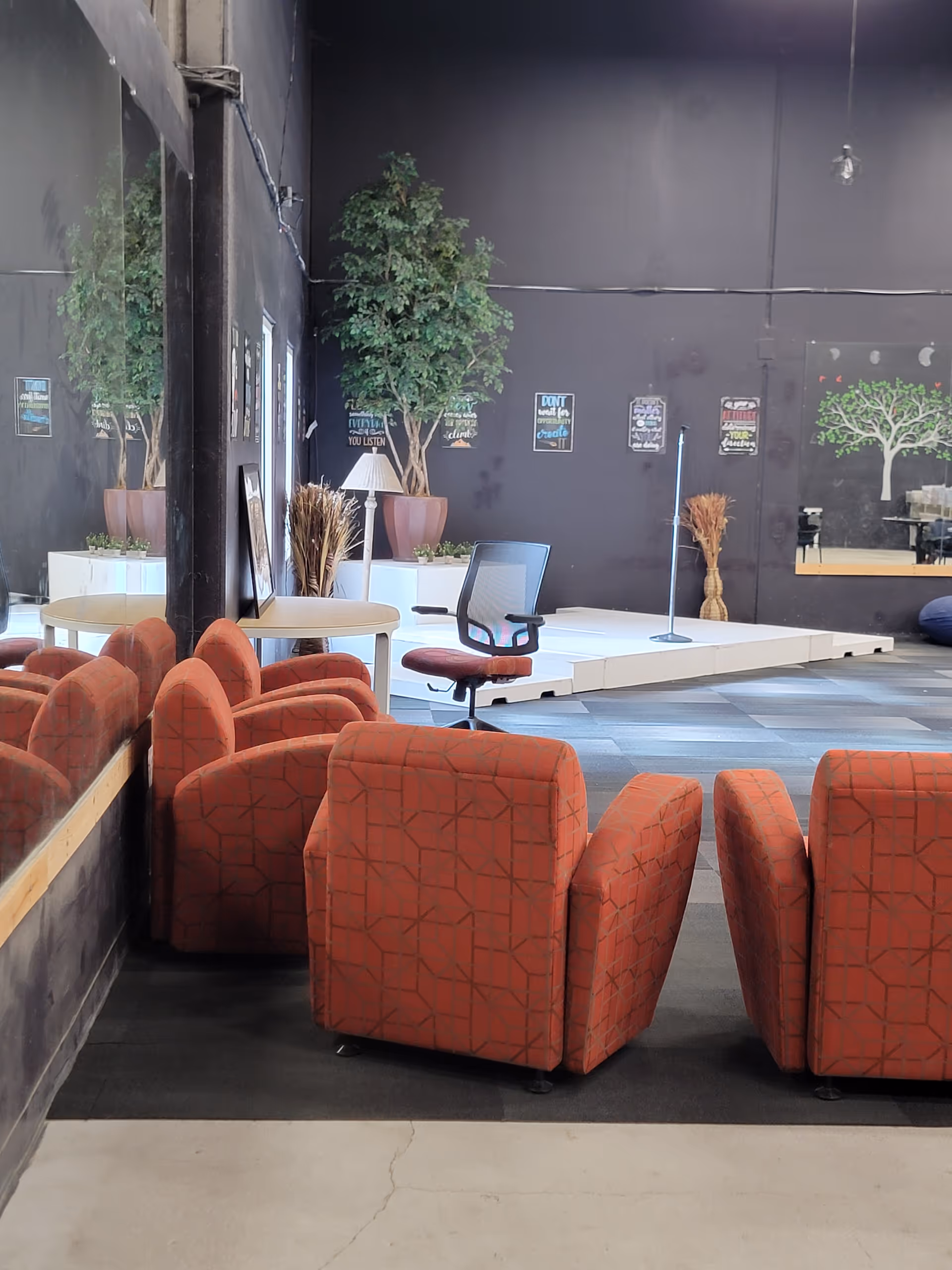 Indoor lounge area with red chairs, plants, and a small stage against dark walls.