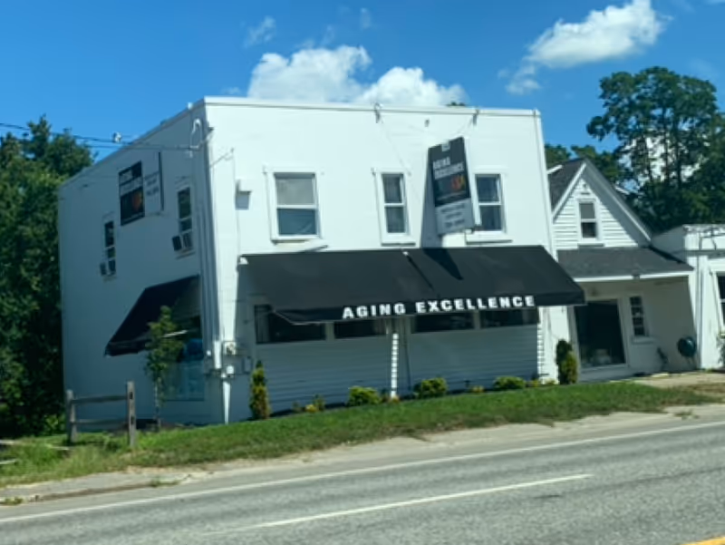 White two-story building with a black awning labeled 'AGING EXCELLENCE' along a street.