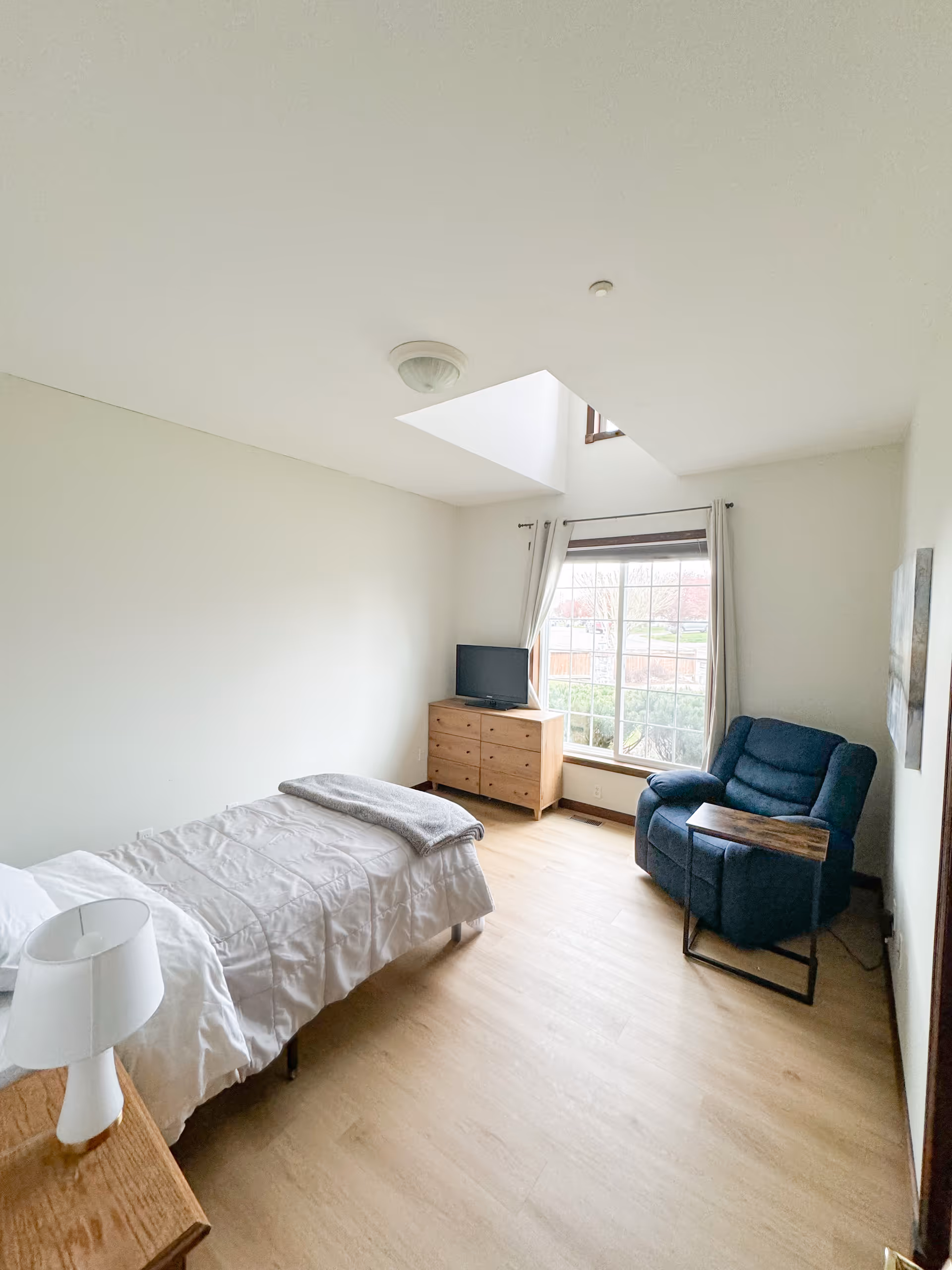 A bright bedroom with a single bed, dresser, TV, chair, and window view.