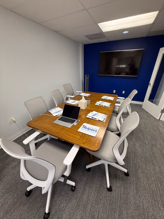A meeting room with a wooden table, chairs, laptop, and wall-mounted TV.