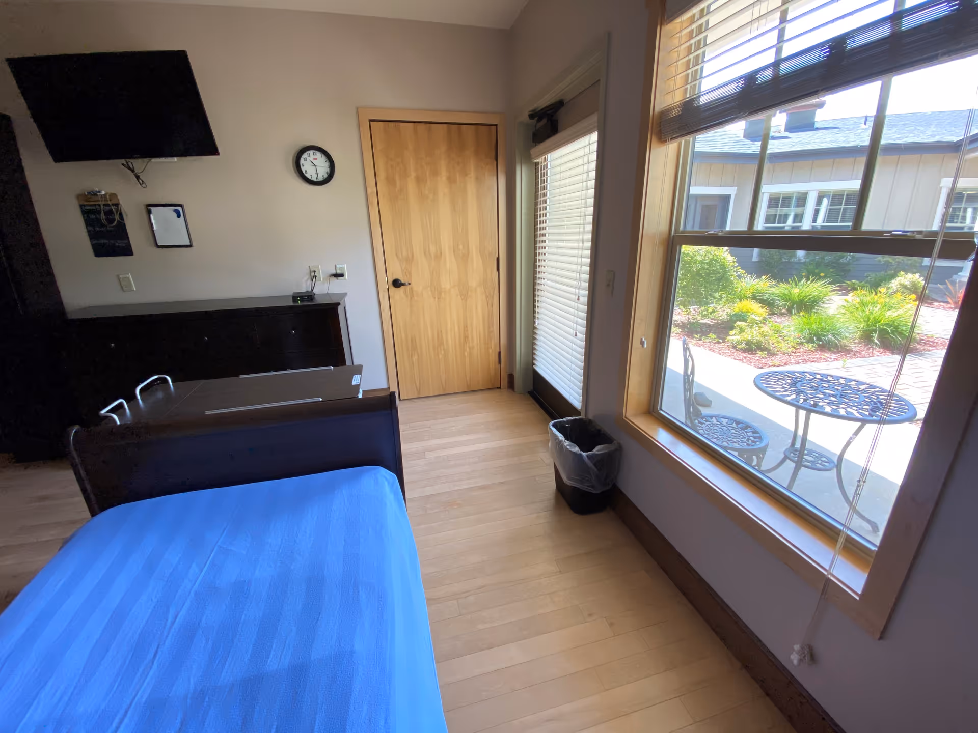 A tidy bedroom with a single bed, dresser, wall clock, and large window view.