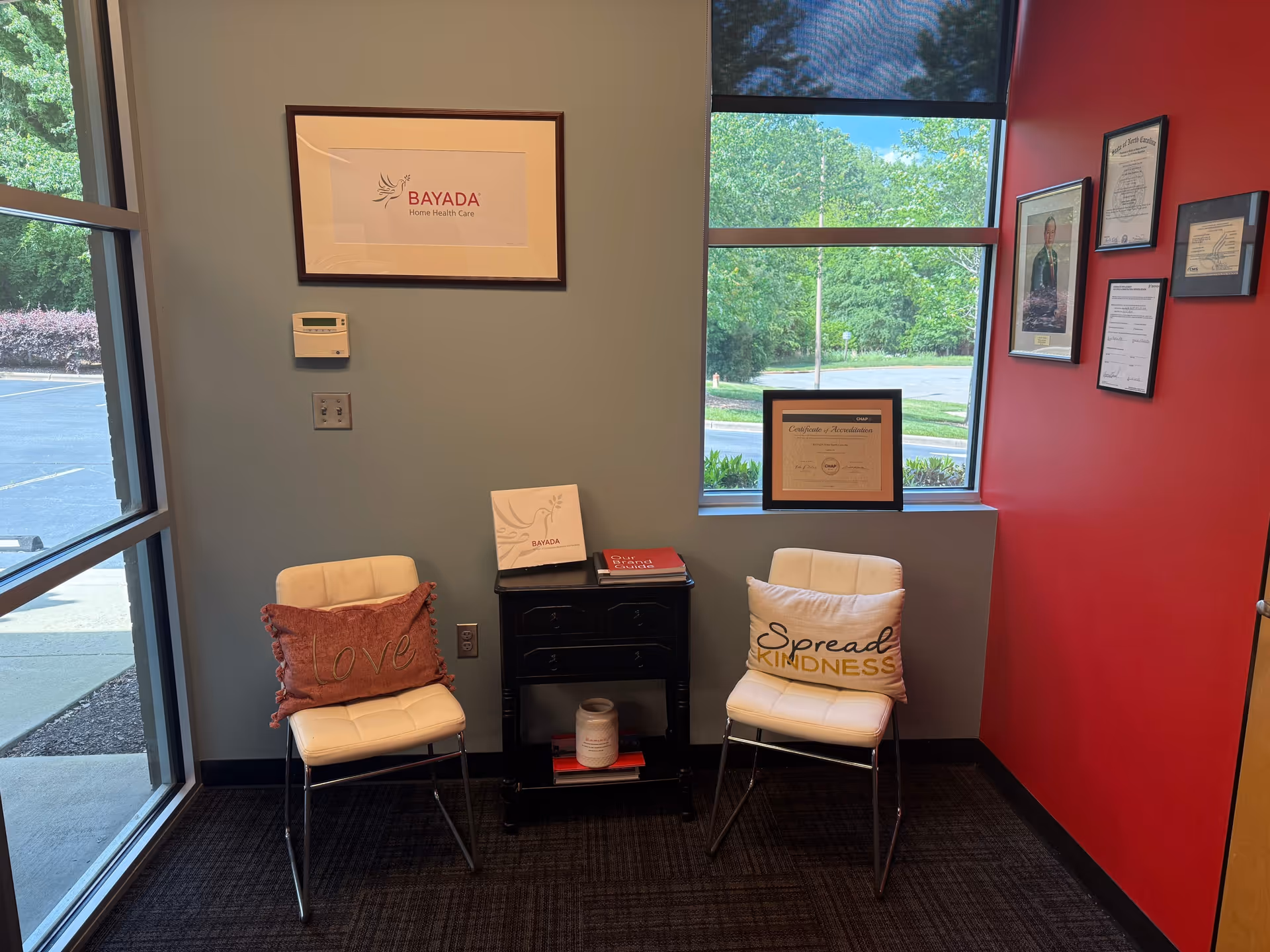 Two white chairs with pillows sit by a window in a BAYADA lobby area.