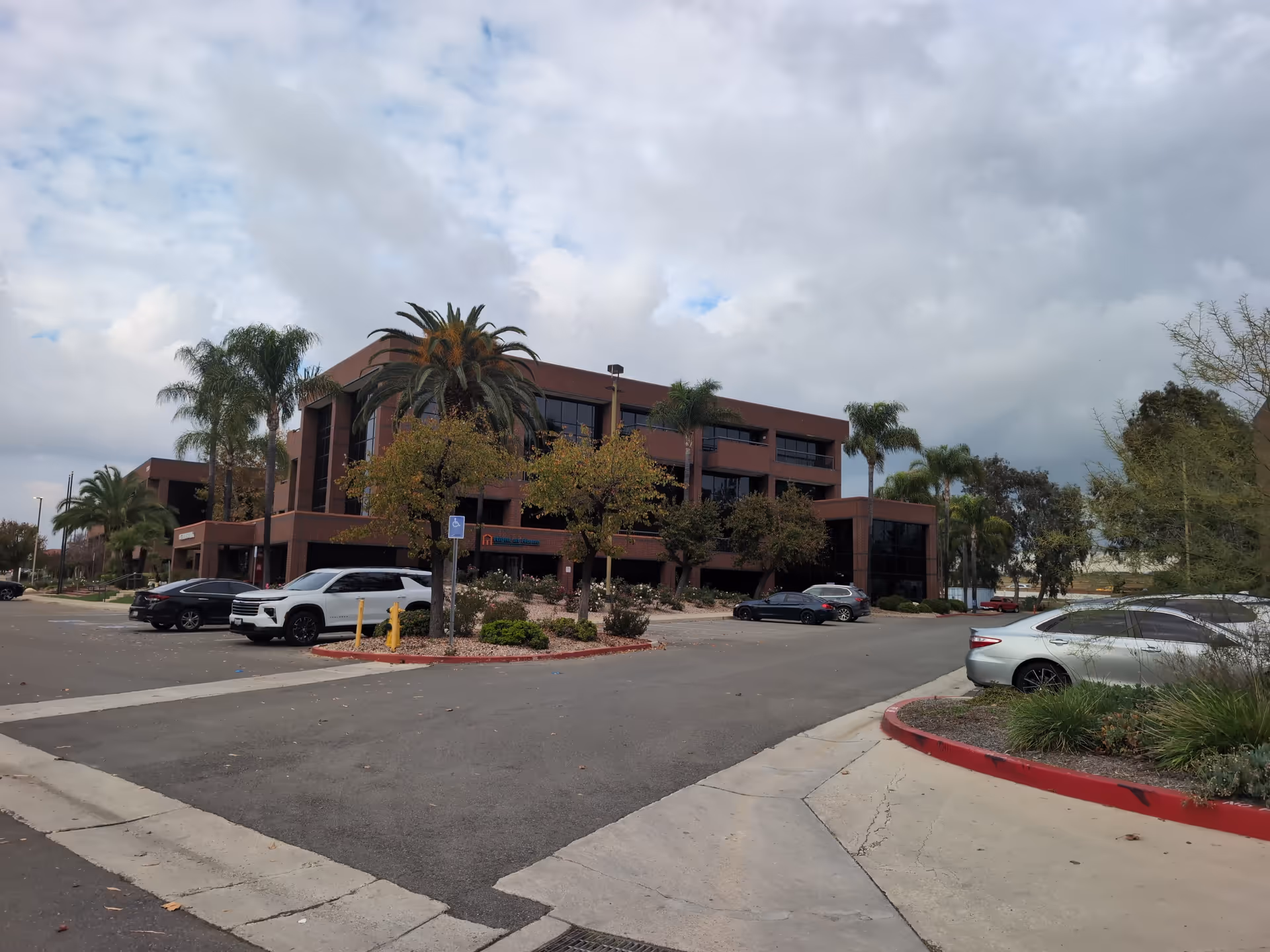 Front view of a brown-orange senior living building with palm trees and parked cars.