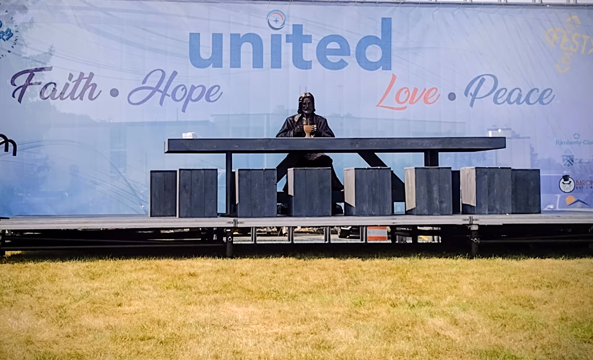 Outdoor stage with a performer seated at a long table in front of a large banner.