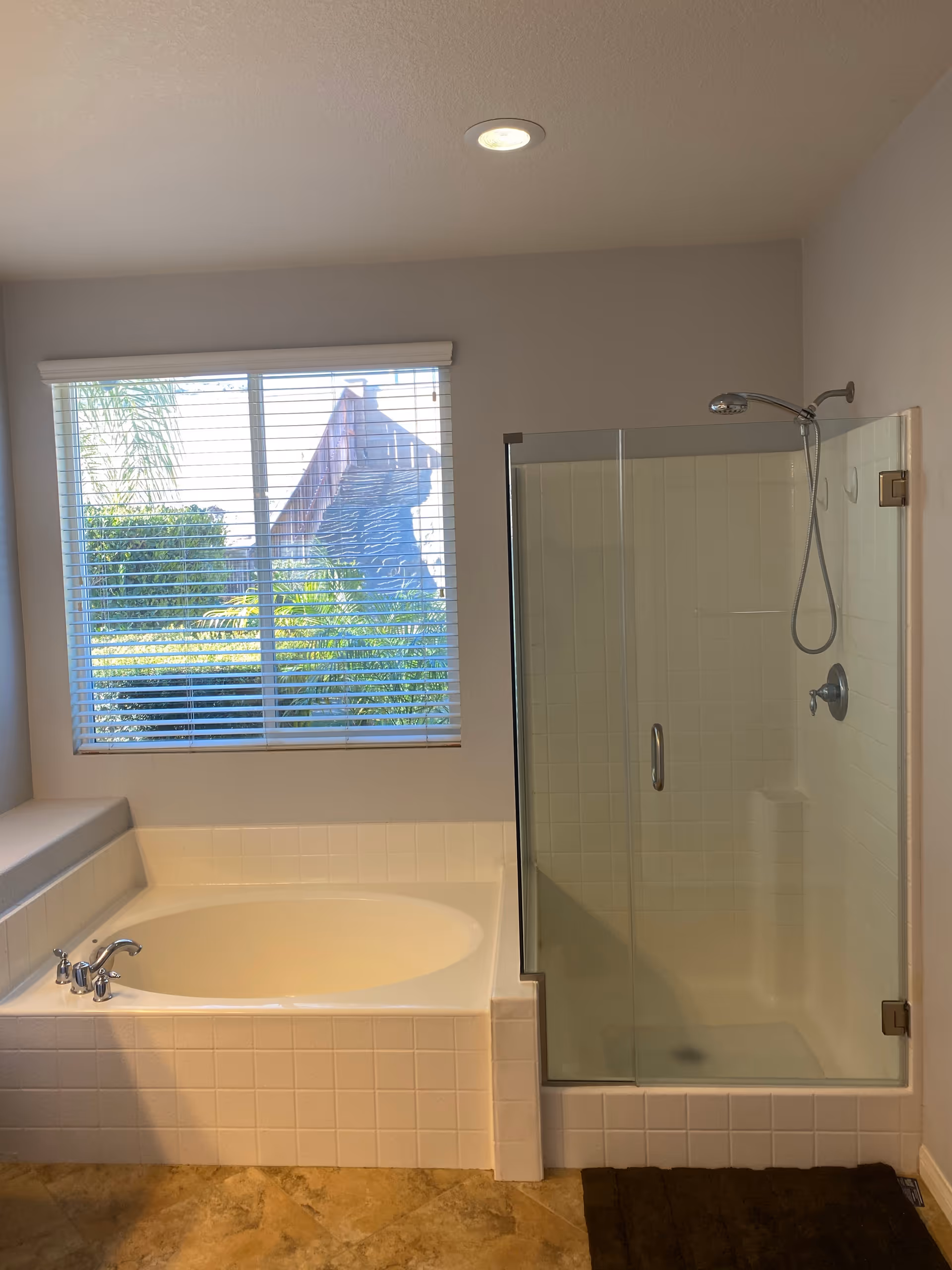 Bright bathroom featuring a tiled tub, glass-enclosed shower, and a window with blinds.