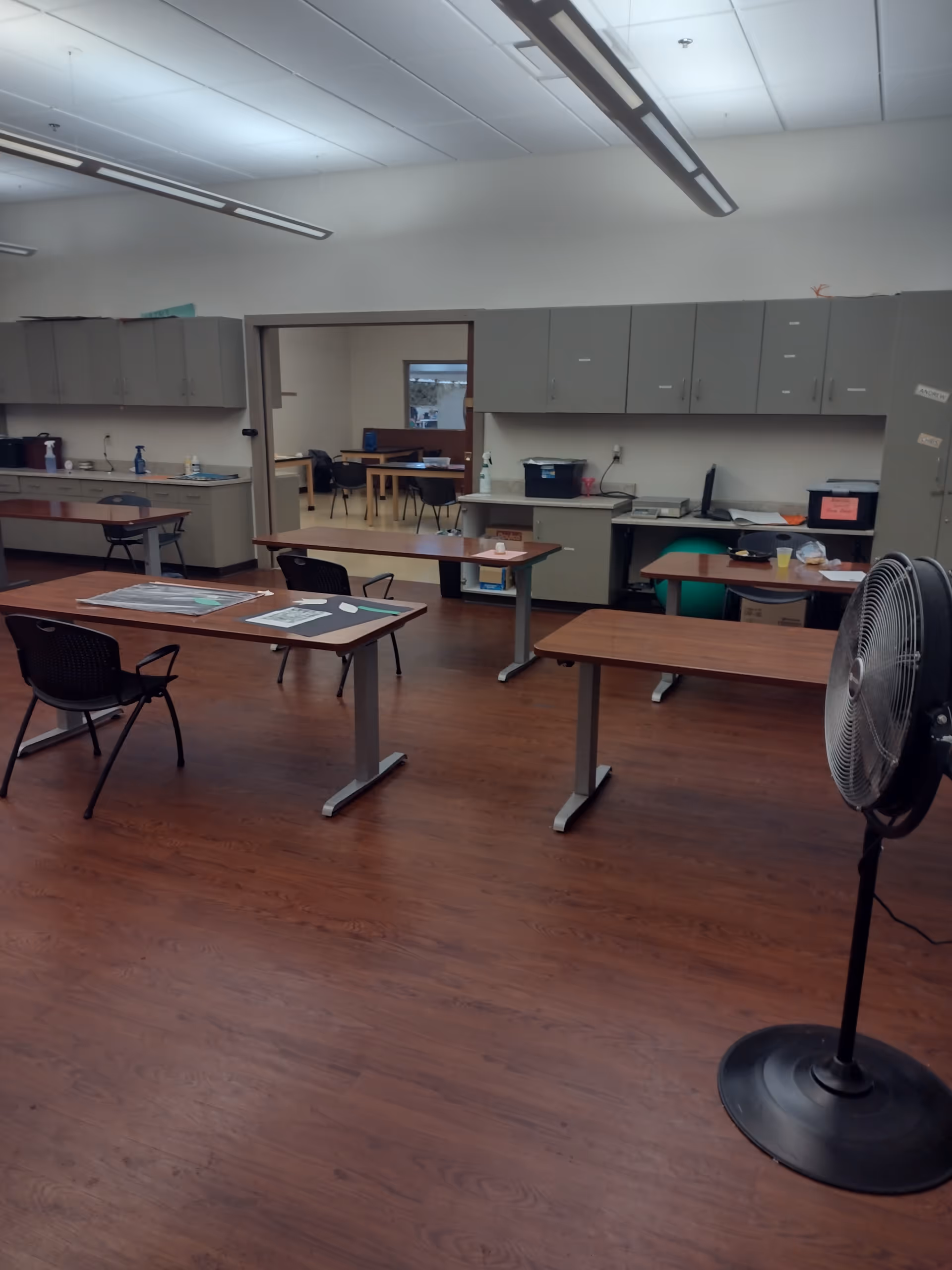 A spacious interior room with tables, chairs, wall cabinets, and a large standing fan.