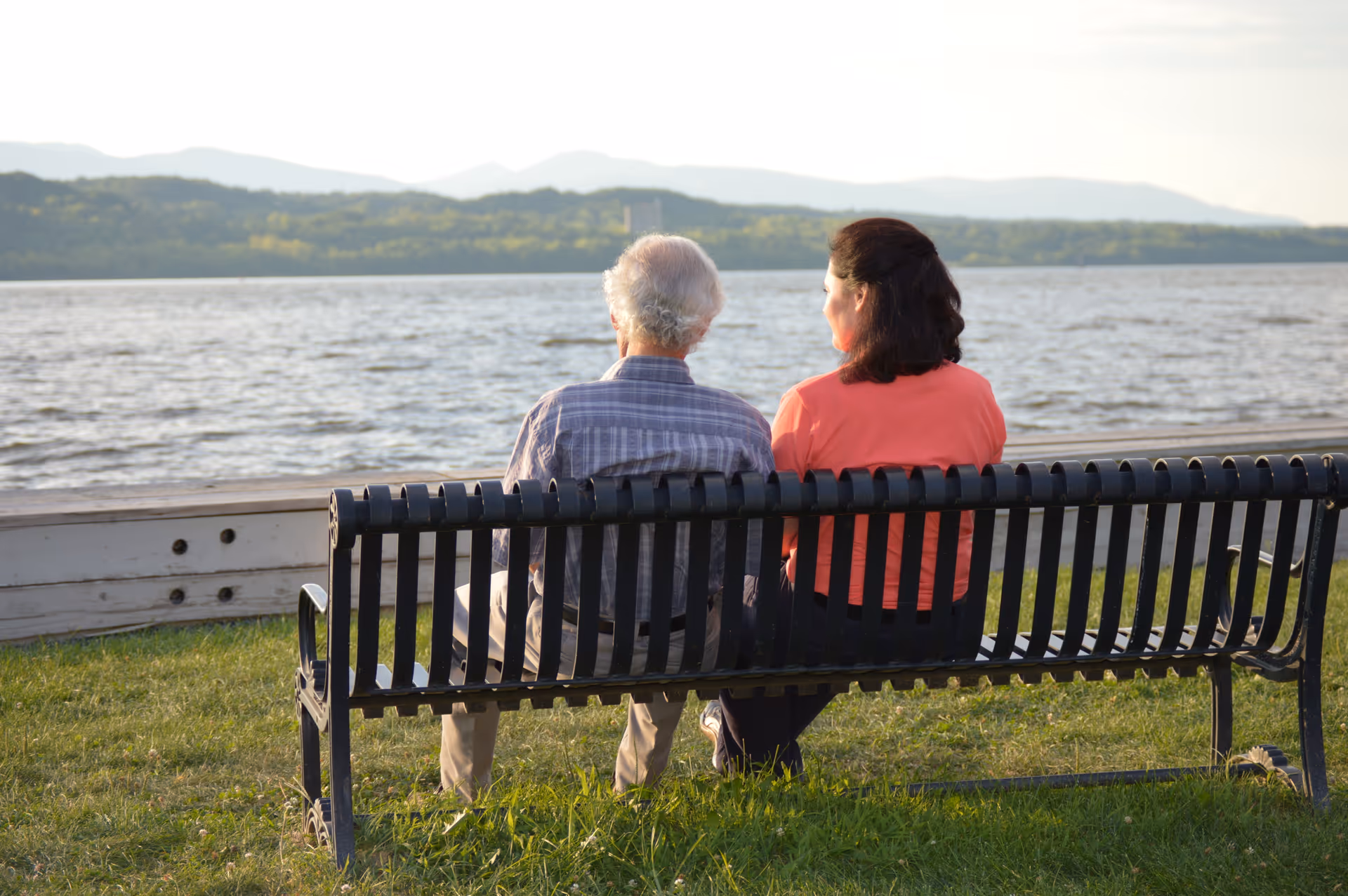 Two elderly individuals sit on a bench by a lake, viewing distant hills.