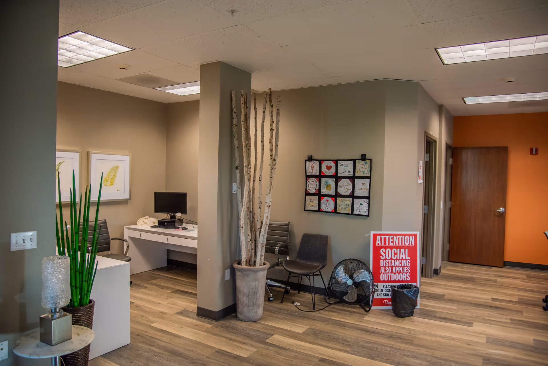 Indoor lobby area with a desk, chairs, plants, and a social distancing sign.