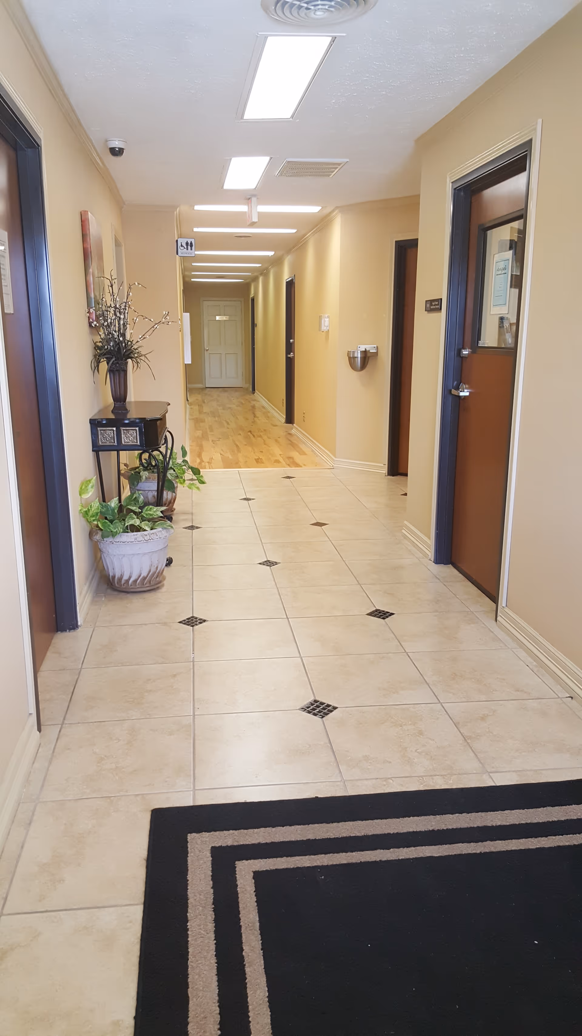 Long beige hallway in a senior living facility with doors, plants, and overhead lights.