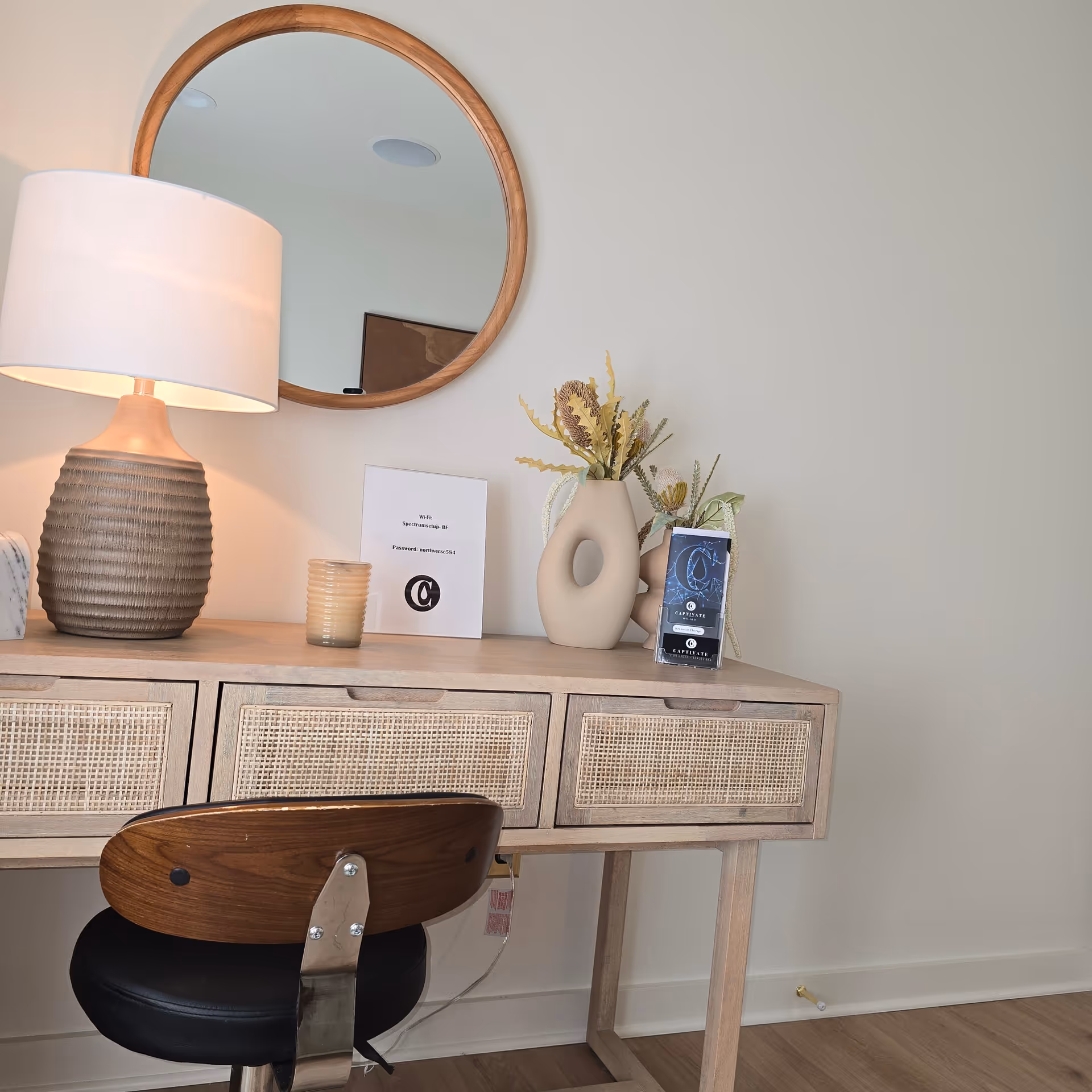 A wooden vanity with a round mirror, lamp, and decorative vases.