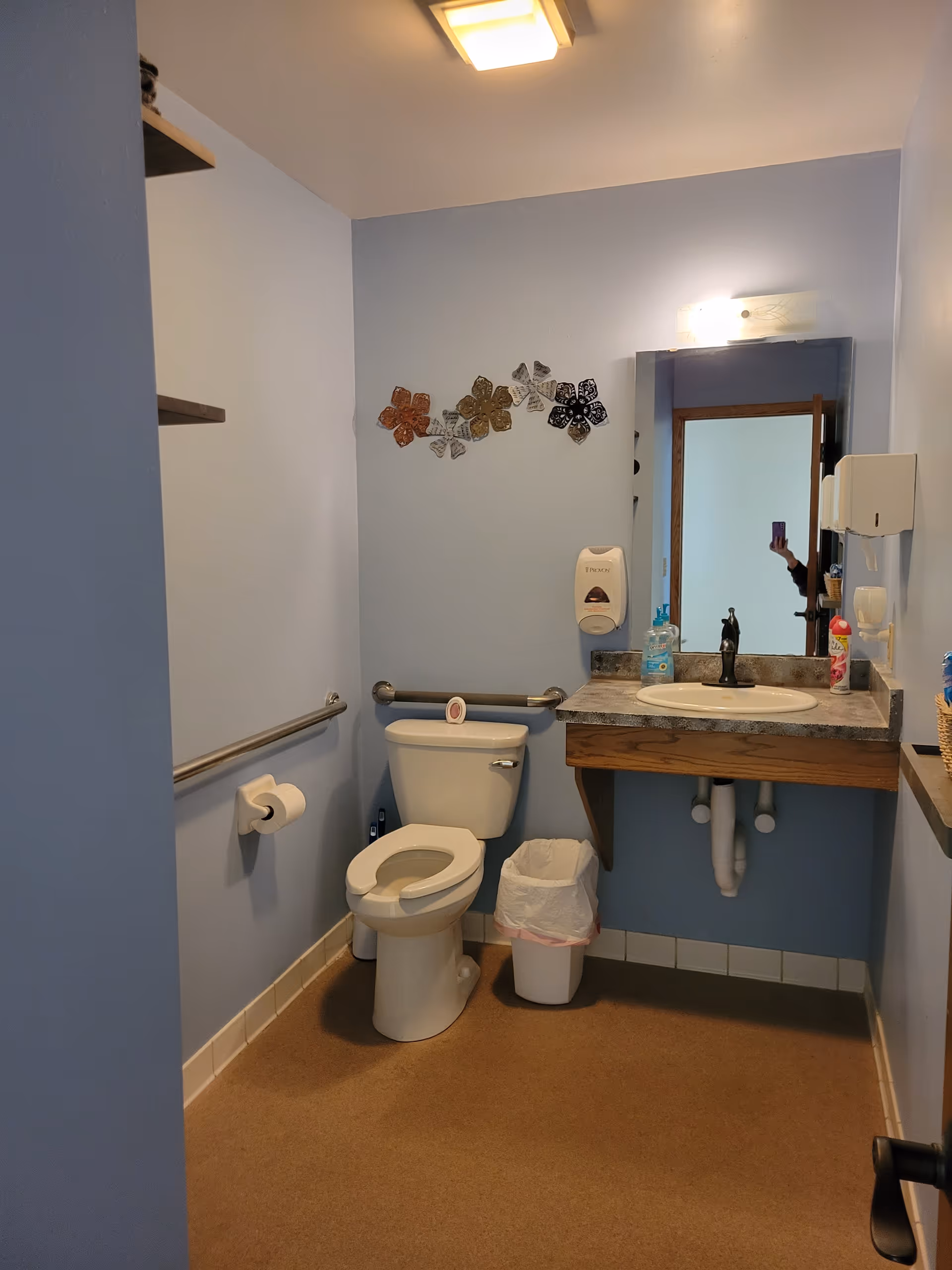 Accessible bathroom with toilet, sink, mirror, grab bars along walls, and trash bin.