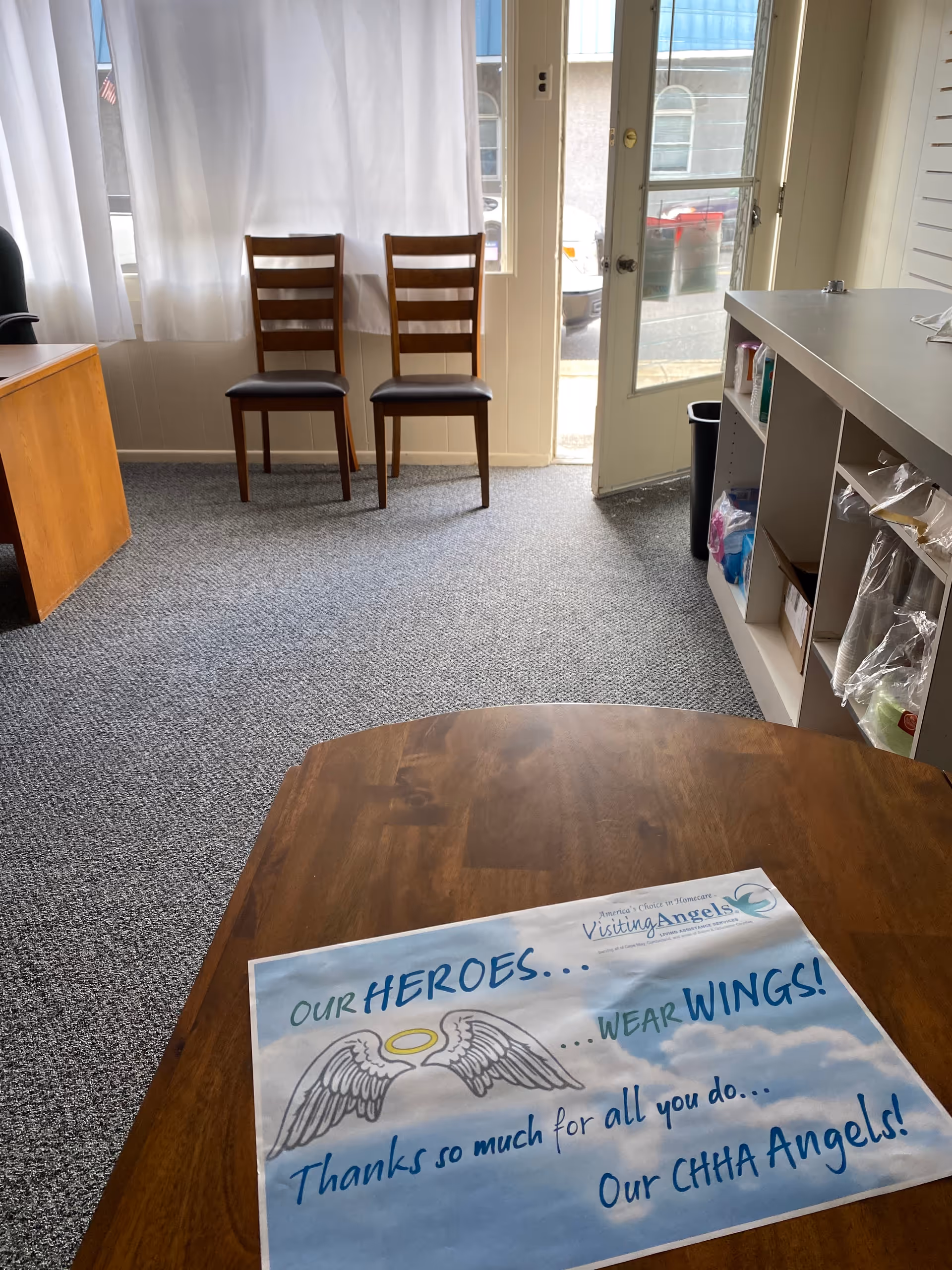 Two wooden chairs sit near windows in a small reception area with a table and poster.