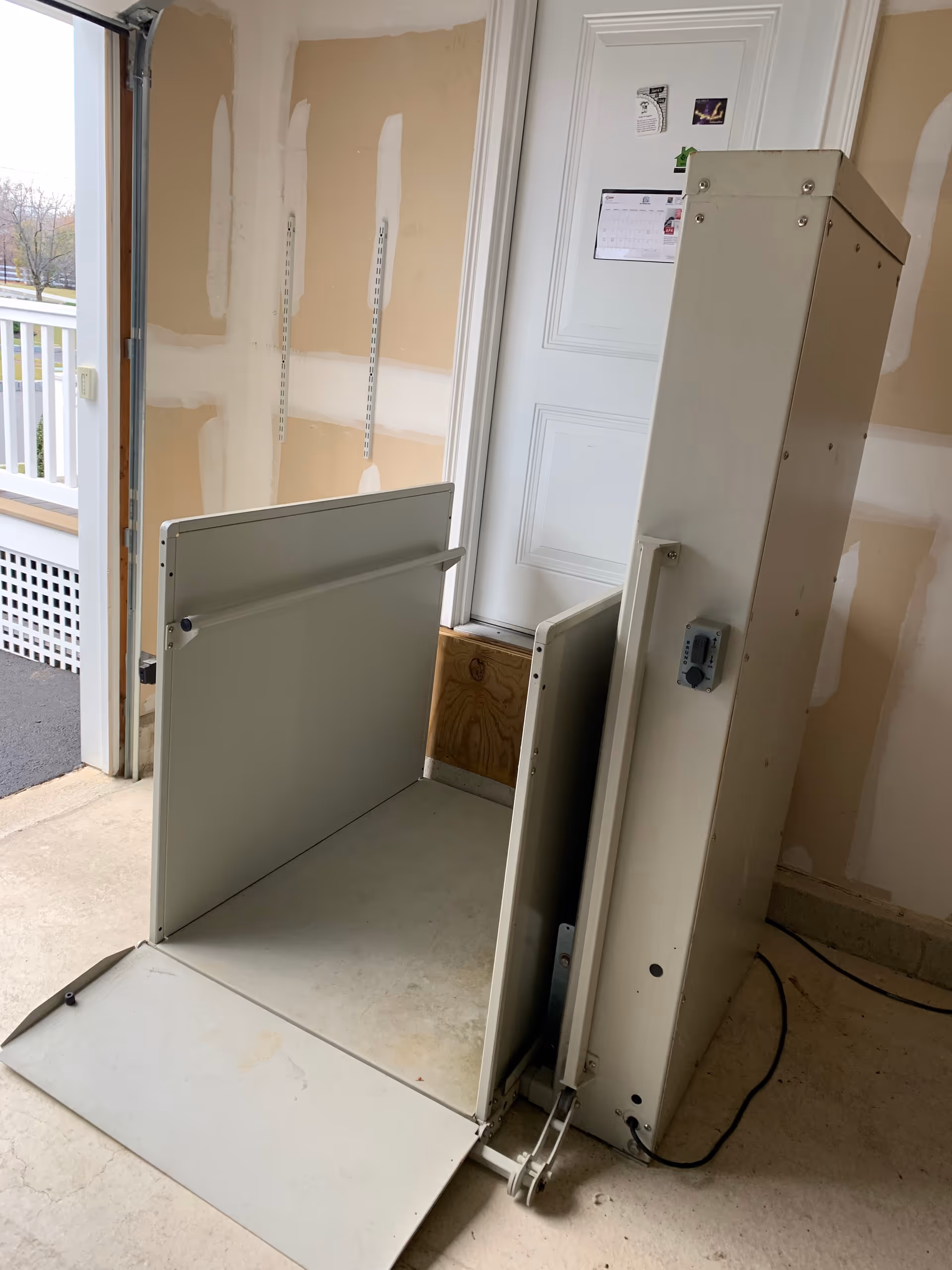 Interior view of a metal platform lift near a doorway in an unfinished hallway.