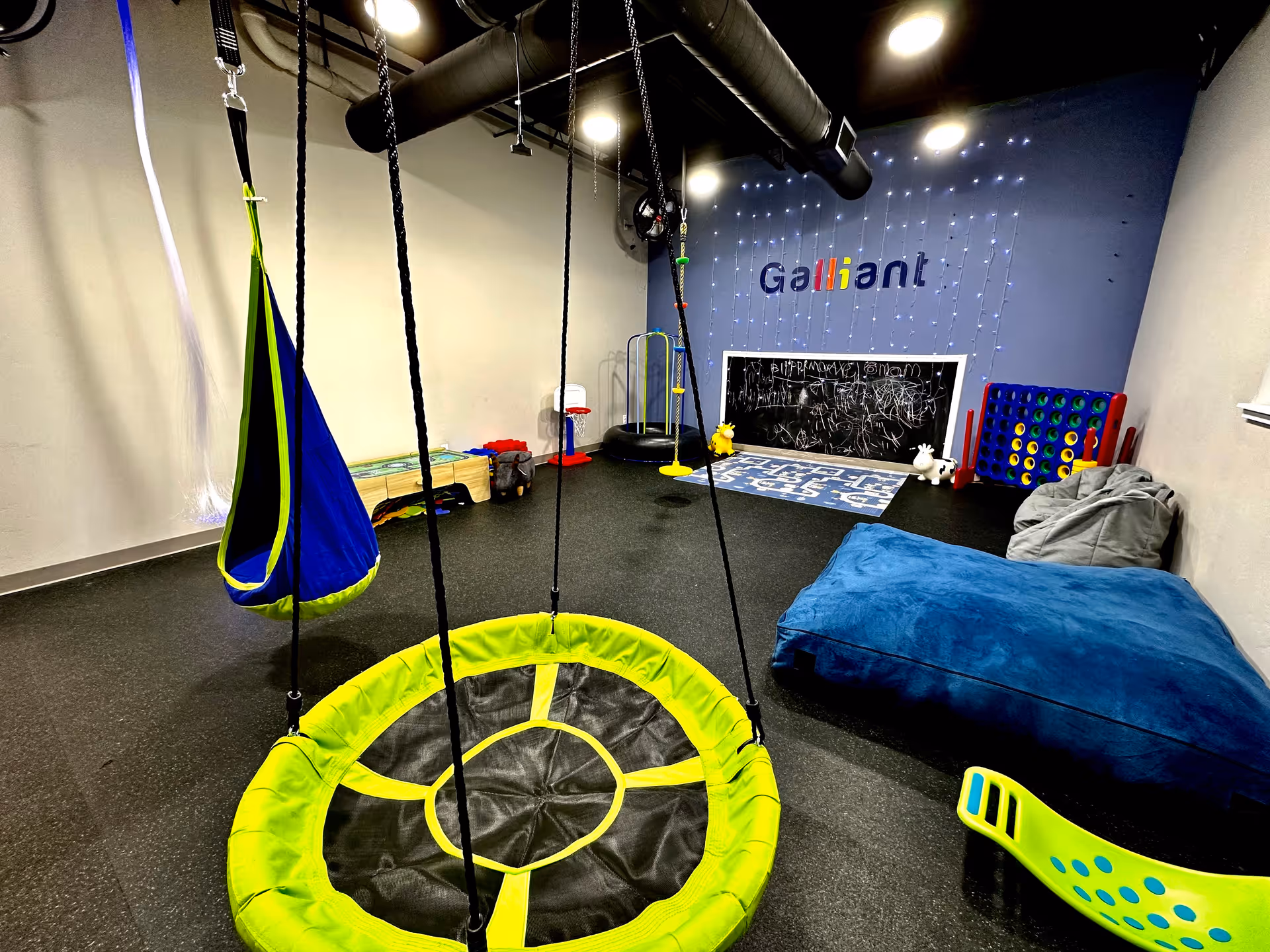 Interior therapy room with swings, a bright circular trampoline, bean bags, and a chalkboard wall.
