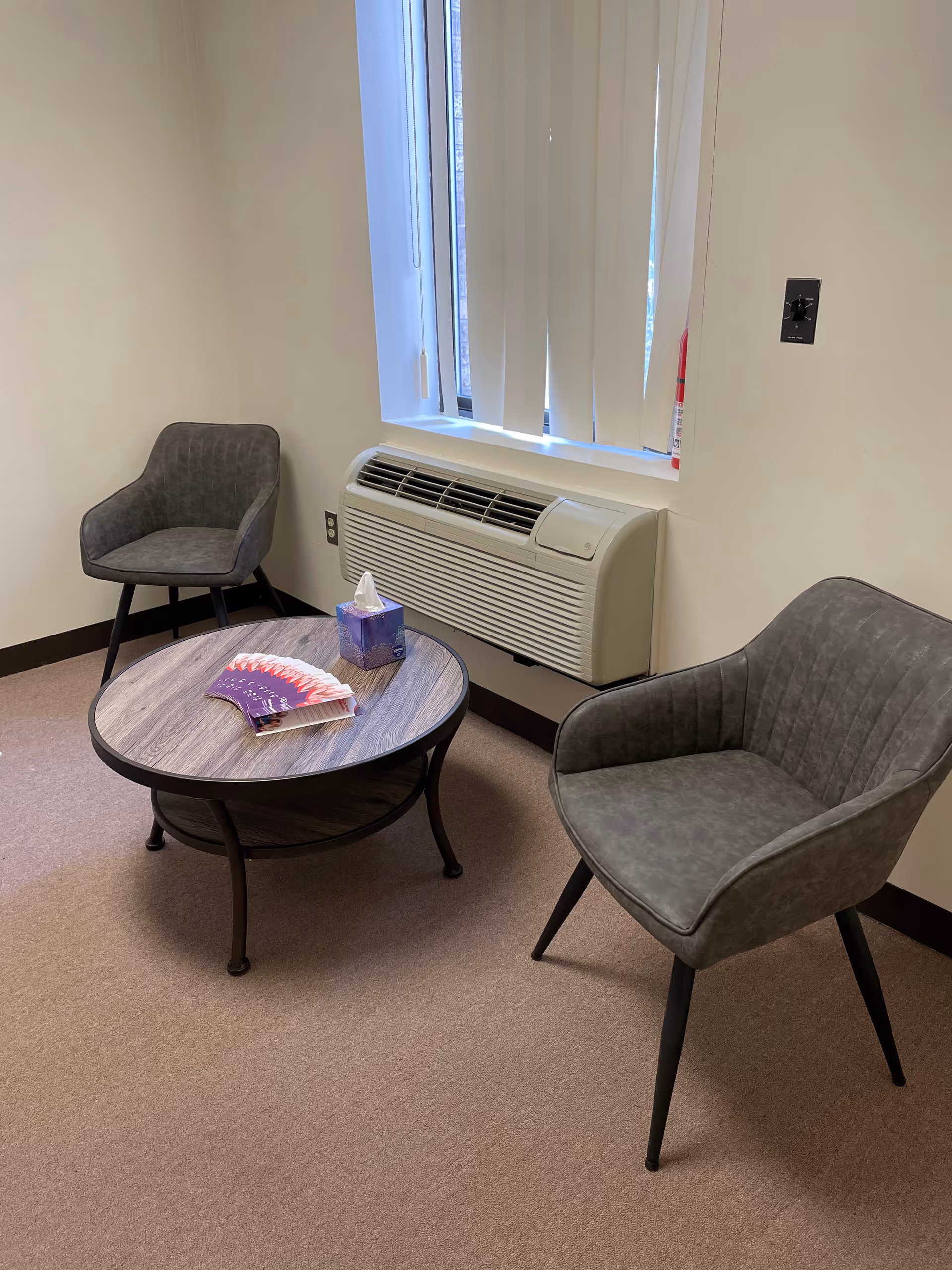 Small waiting area with two gray chairs around a round table near a window.