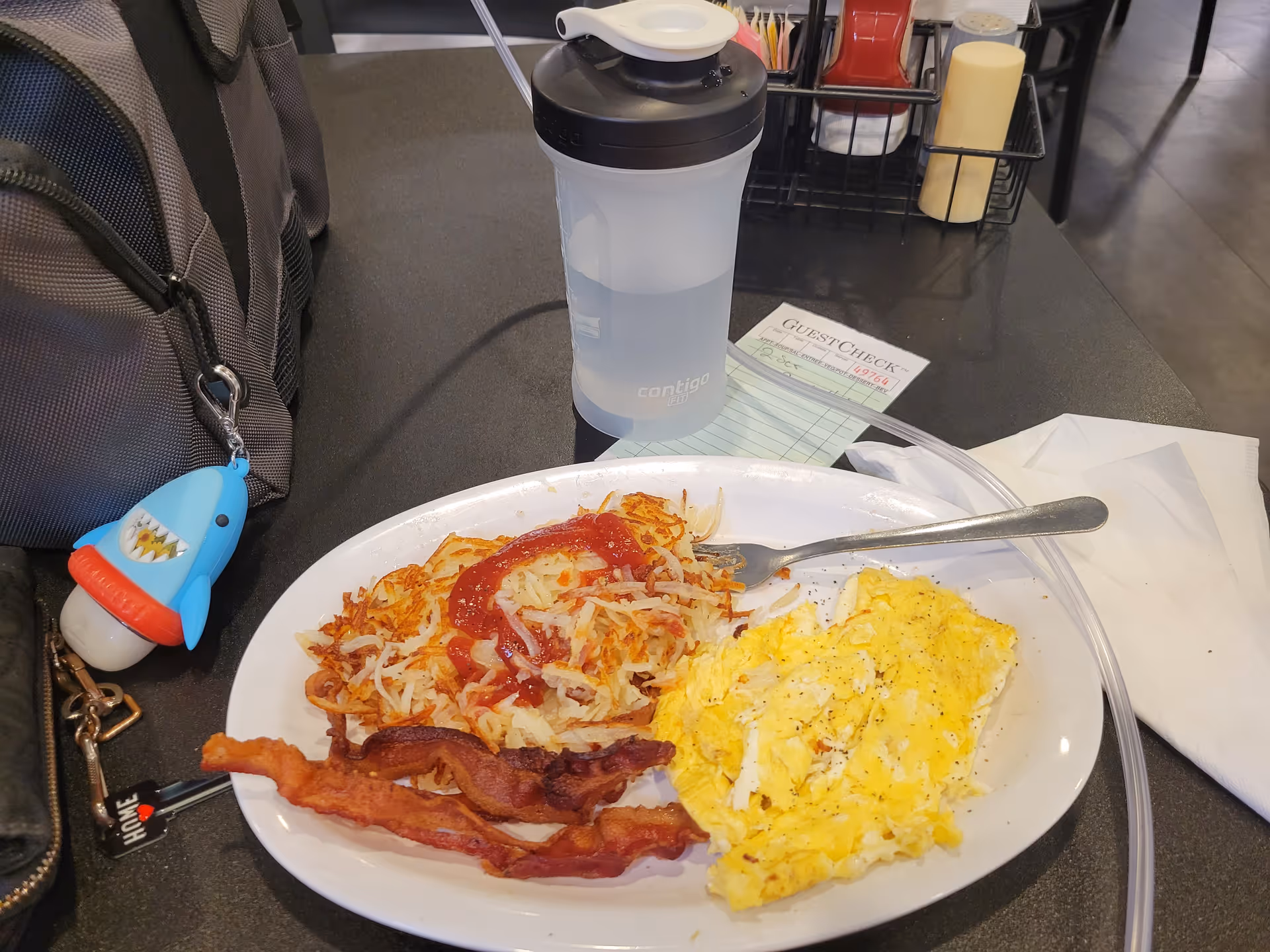 Plate with scrambled eggs, hash browns, and bacon on a table beside a cup and condiments.