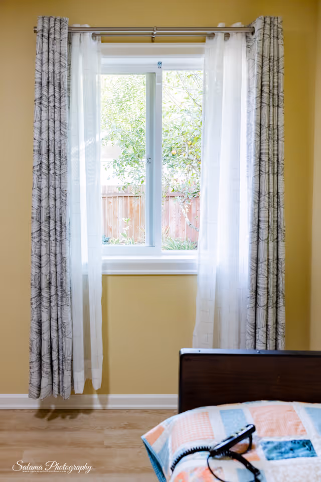Bright yellow bedroom with a large window, sheer curtains, and a portion of a bed.