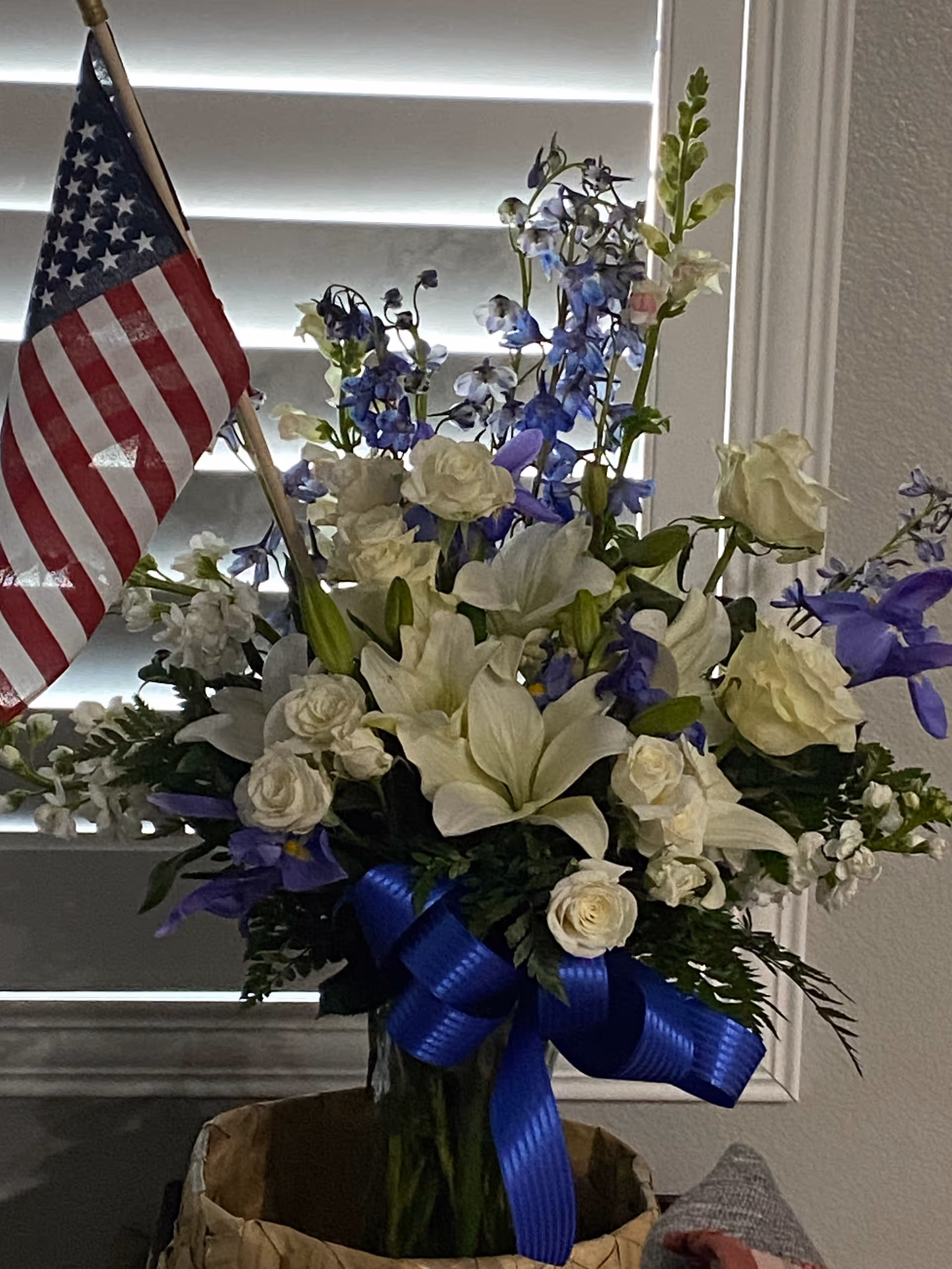 Colorful flower arrangement with white lilies and roses, blue ribbon, and a small American flag indoors.