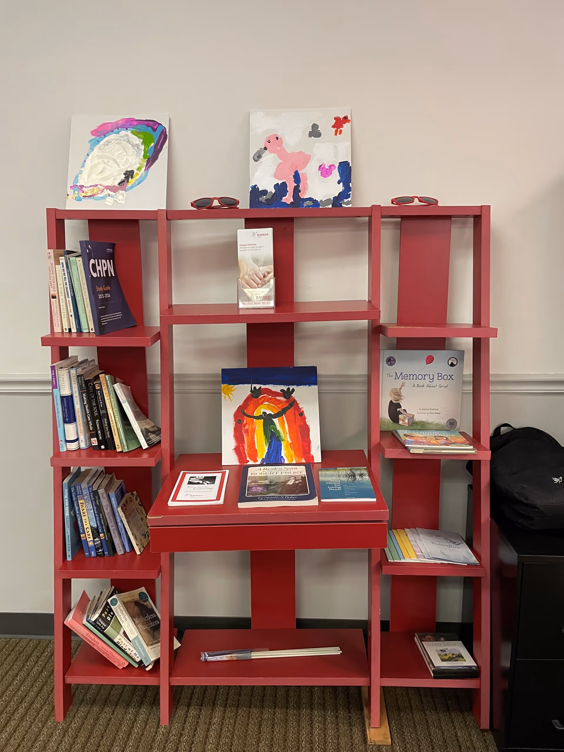 Red bookshelf with books and artwork against a light wall in a quiet interior.