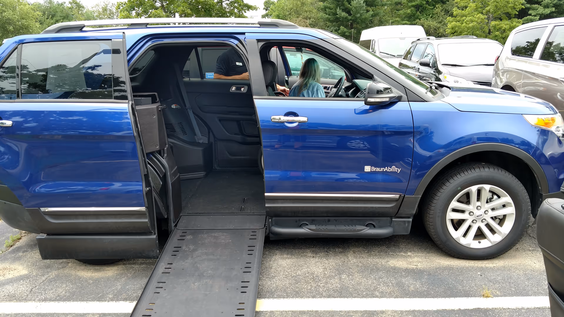 Blue wheelchair-accessible van with ramp extended in parking lot; two people inside.