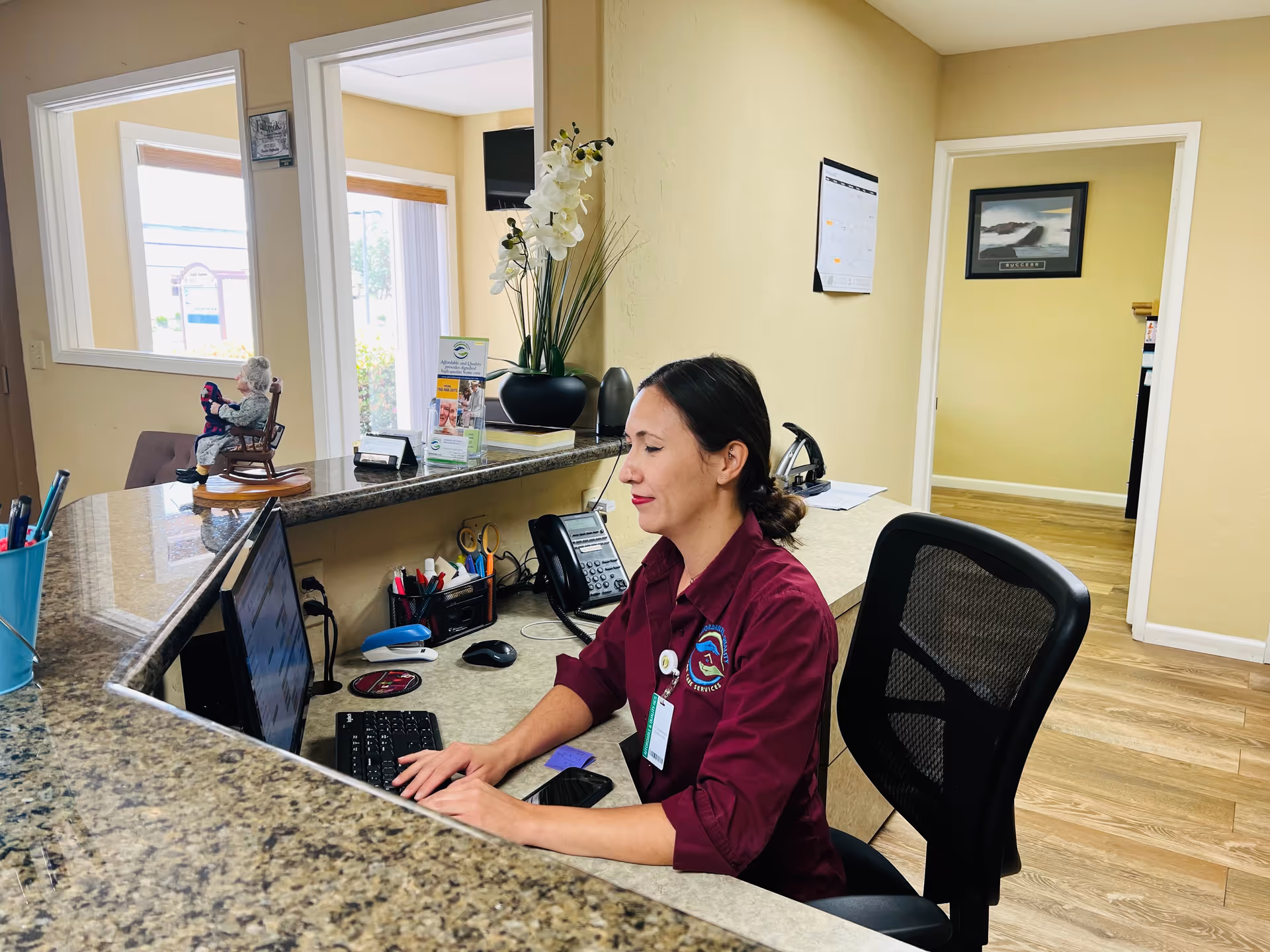 Caregiver at a granite reception desk uses a laptop; resident seated in the background.