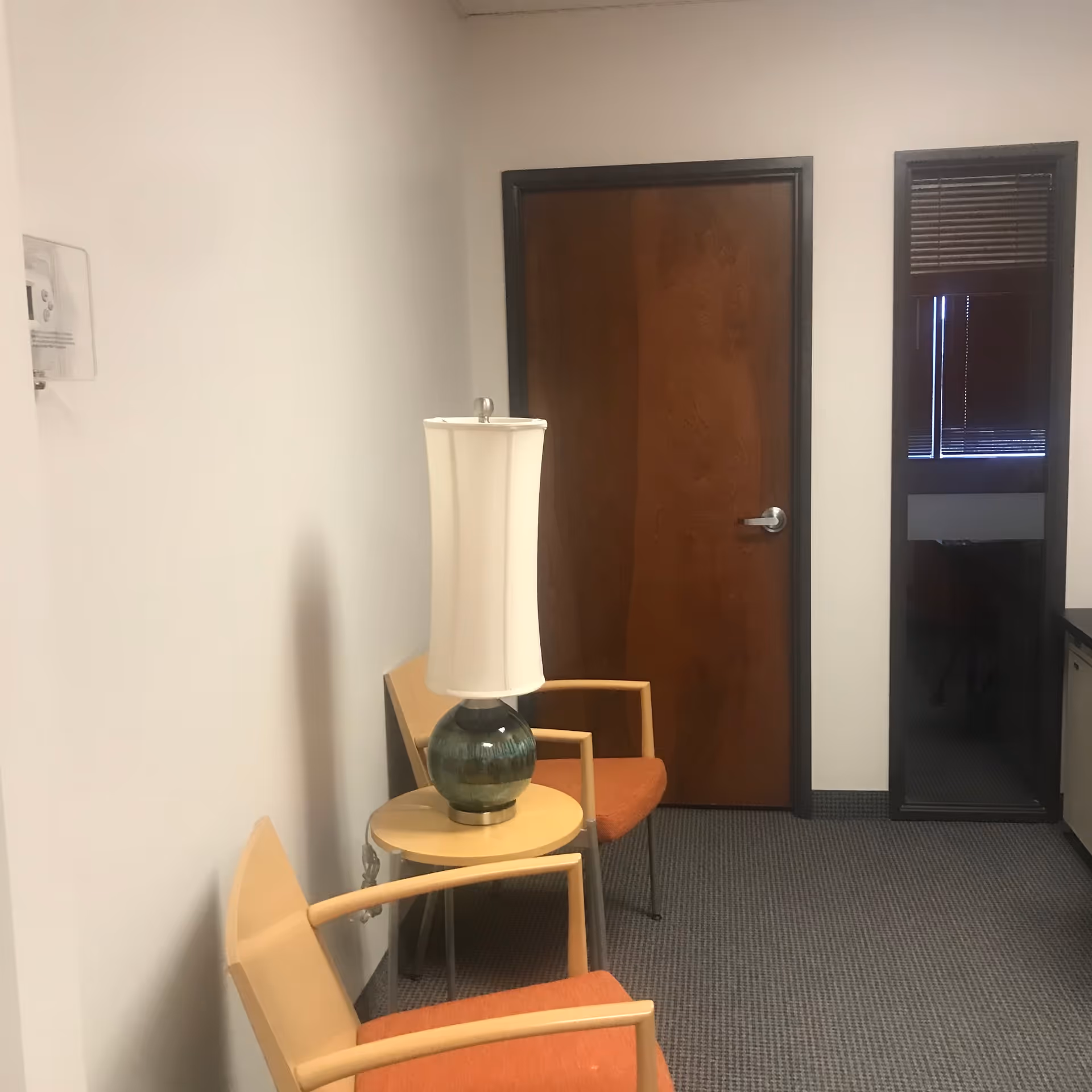 A small waiting area with chairs, a table, and a lamp beside a wooden door.