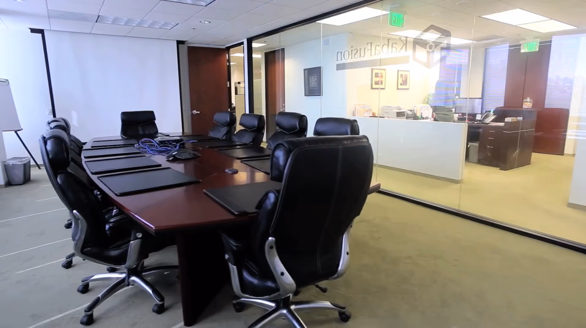 A conference room with a long wooden table, black rolling chairs, and glass-walled offices beyond.