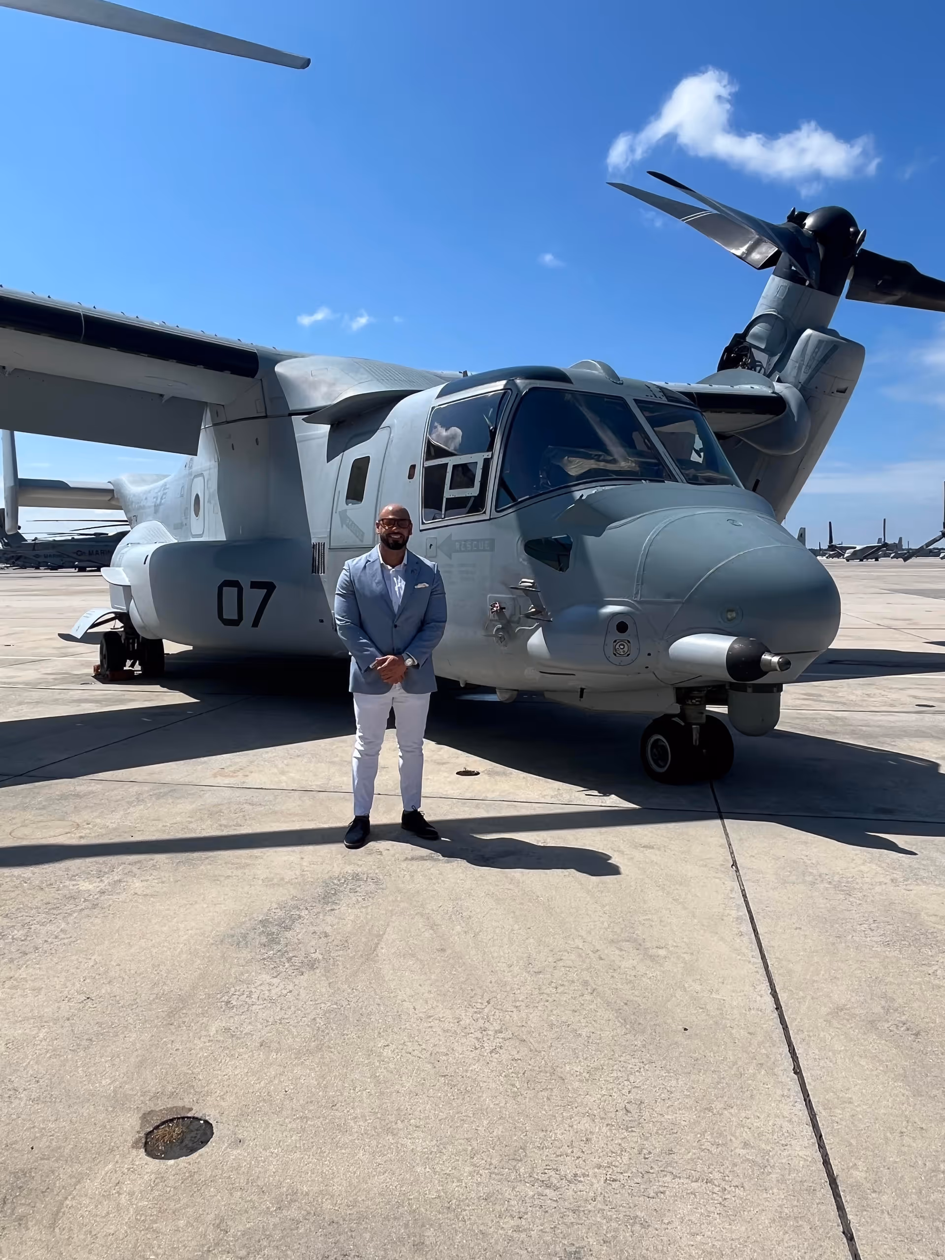 A man in a light blazer stands beside a gray helicopter on a sunny tarmac.