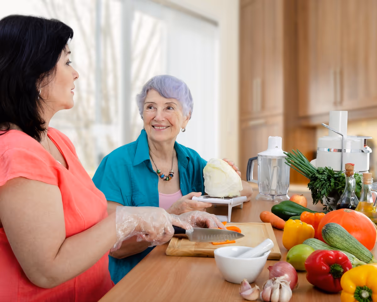 Two women prepare vegetables at a bright kitchen counter with a cutting board.