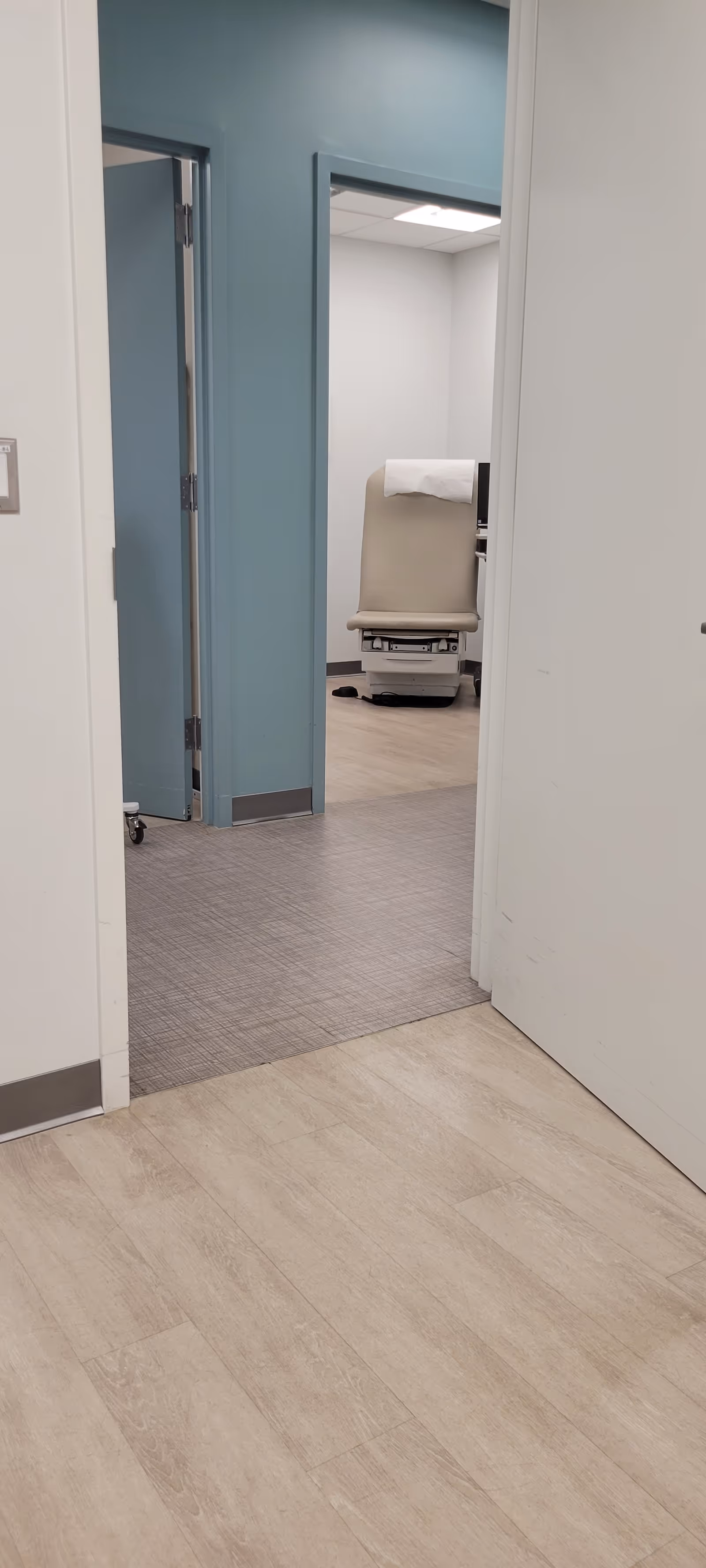 Hallway leads to a room with a beige exam chair in blue-walled clinical space.