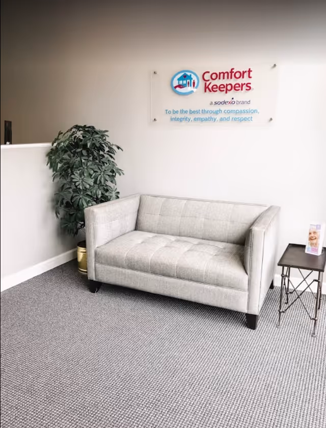 Lobby seating area with a gray sofa, plant, and Comfort Keepers sign.