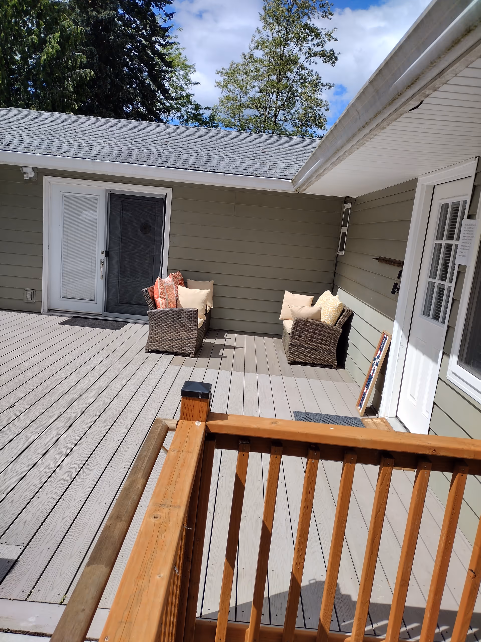 Outdoor wooden deck with wicker chairs and cushions adjacent to a light green building.