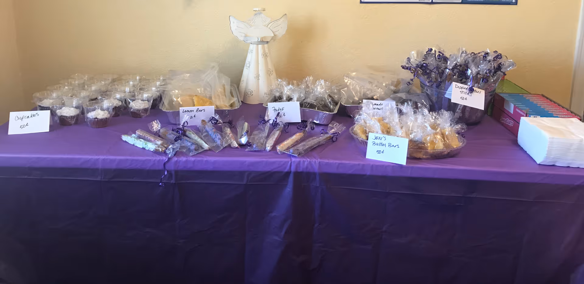 A long table covered with a purple cloth displays wrapped cupcakes and treats indoors.