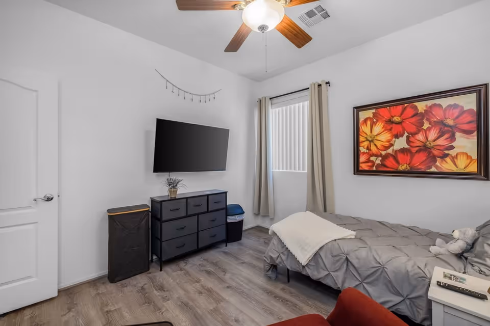 A tidy bedroom with a bed, dresser, wall art, curtains, and a mounted TV.