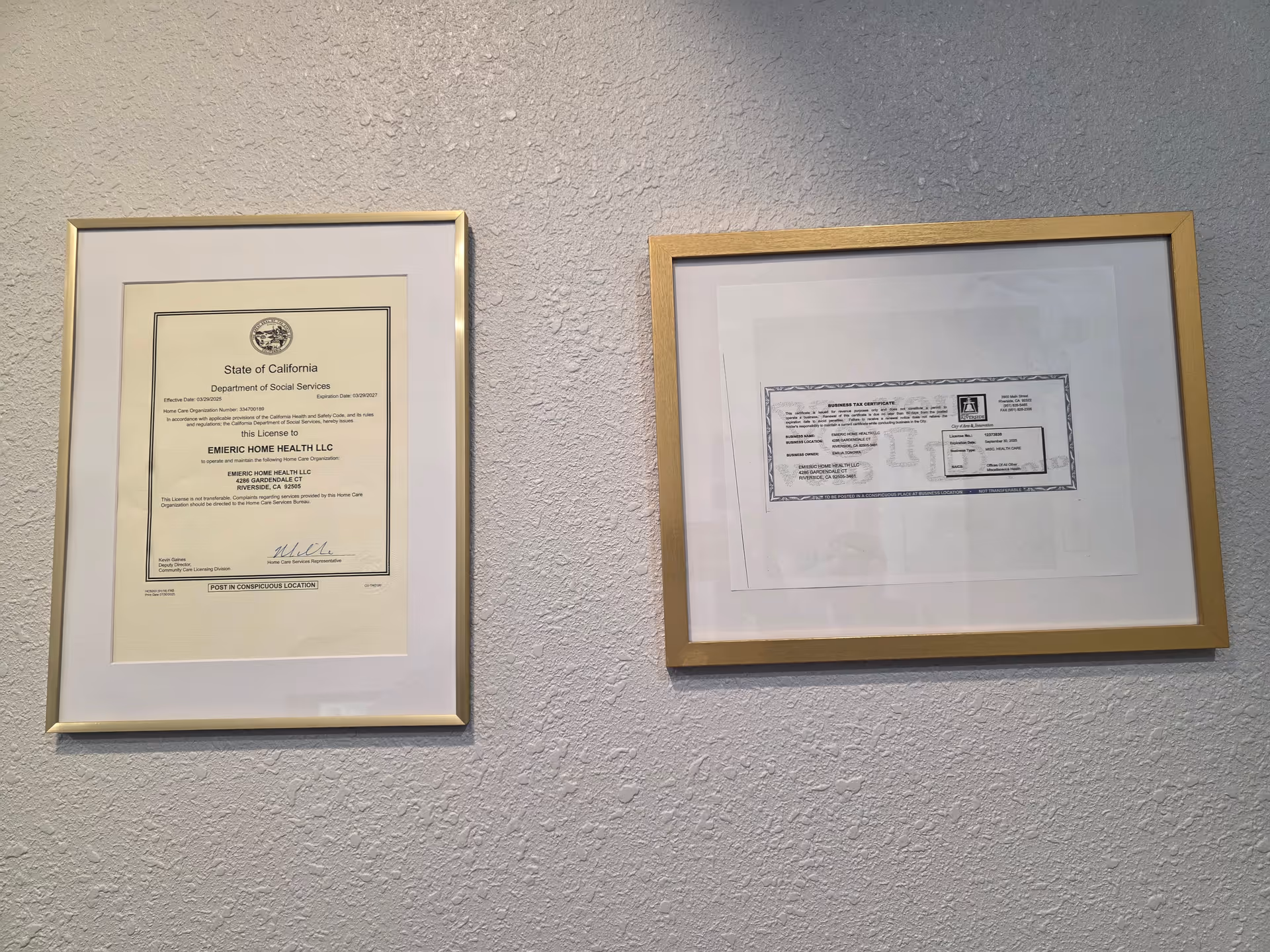 Two framed documents hang on a textured interior wall inside a facility.