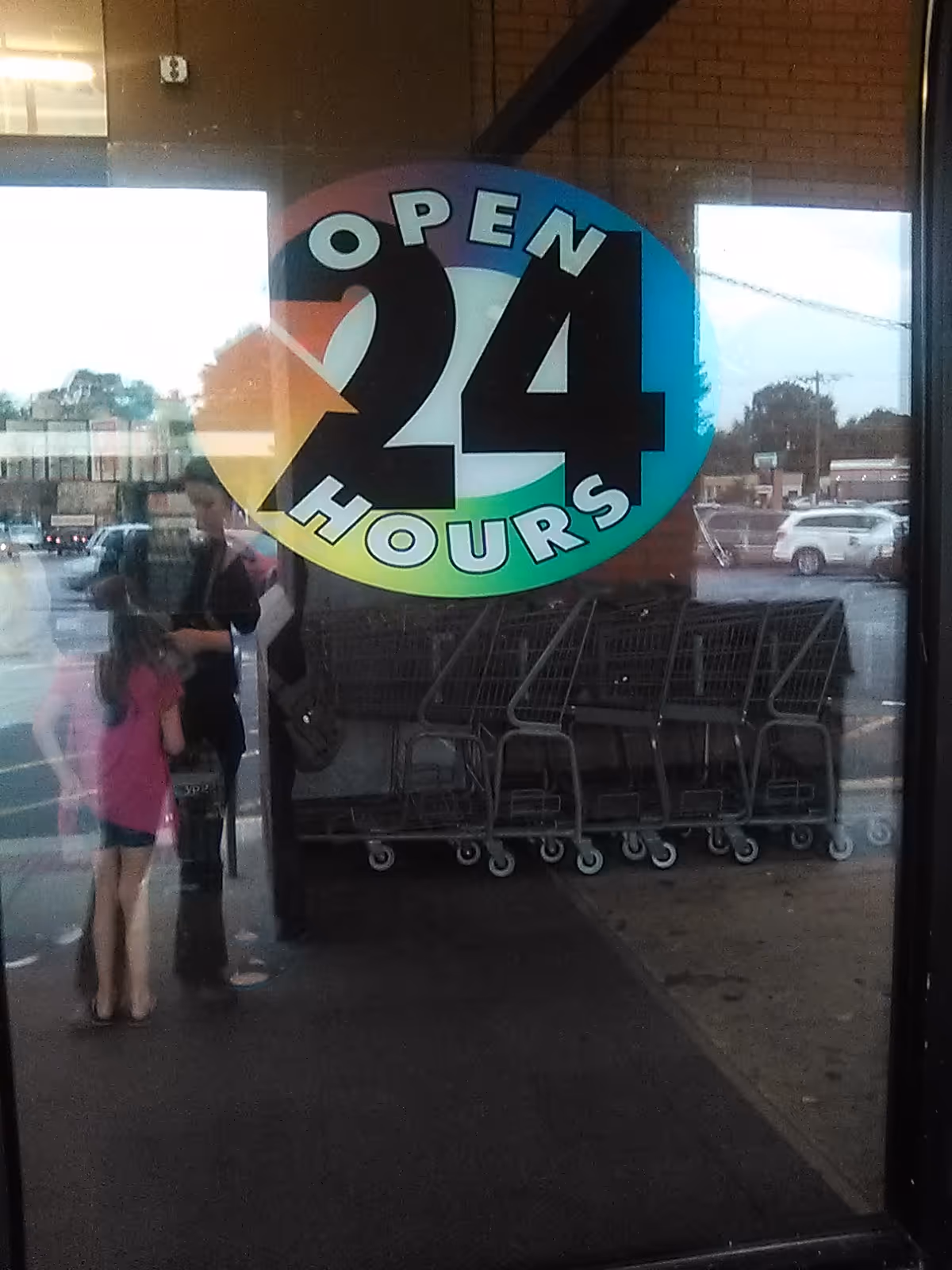 Glass storefront with a colorful OPEN 24 HOURS sticker and shopping carts outside.
