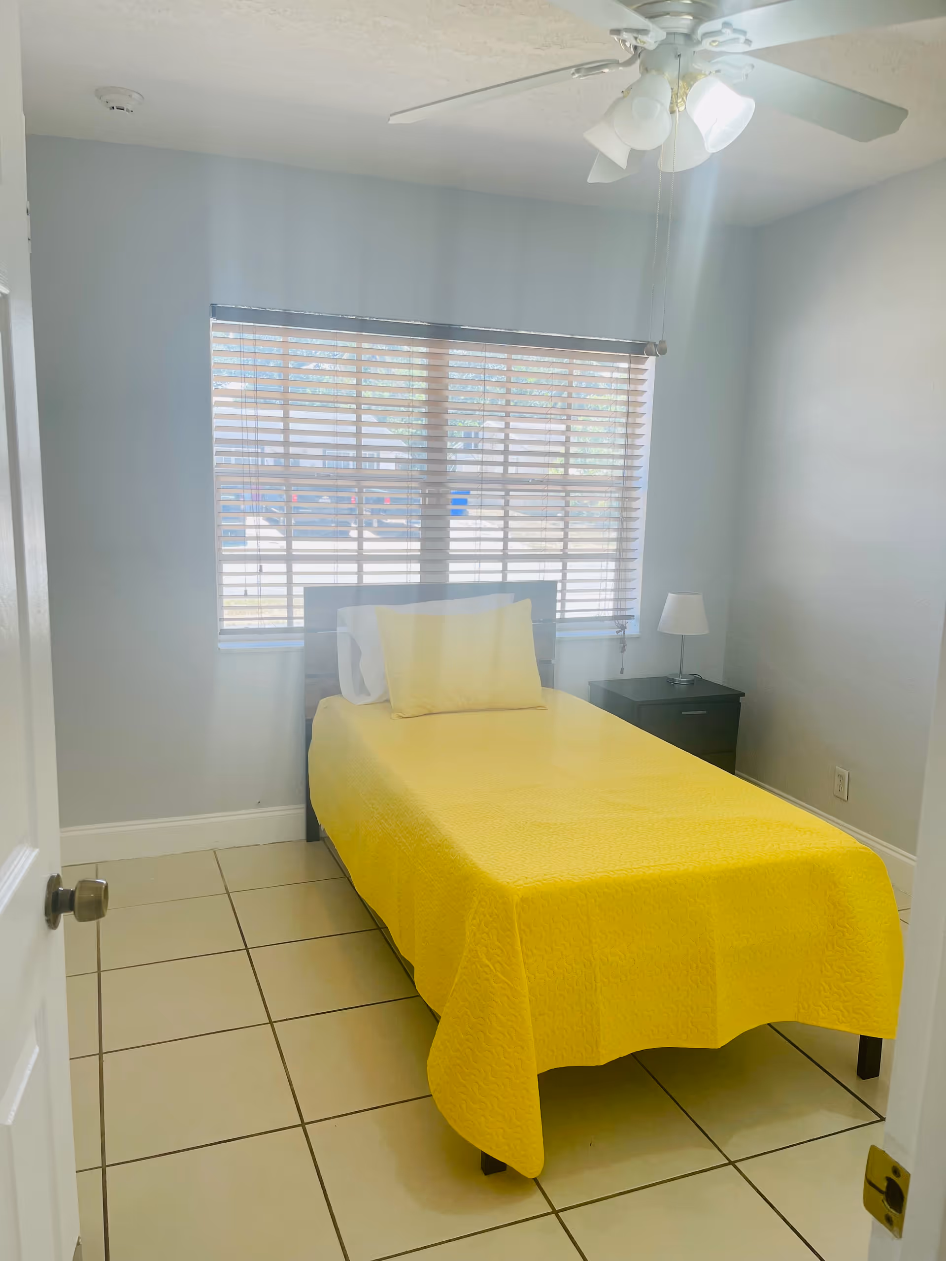 Small bedroom with yellow bedspread, nightstand, lamp, window blinds, and ceiling fan.