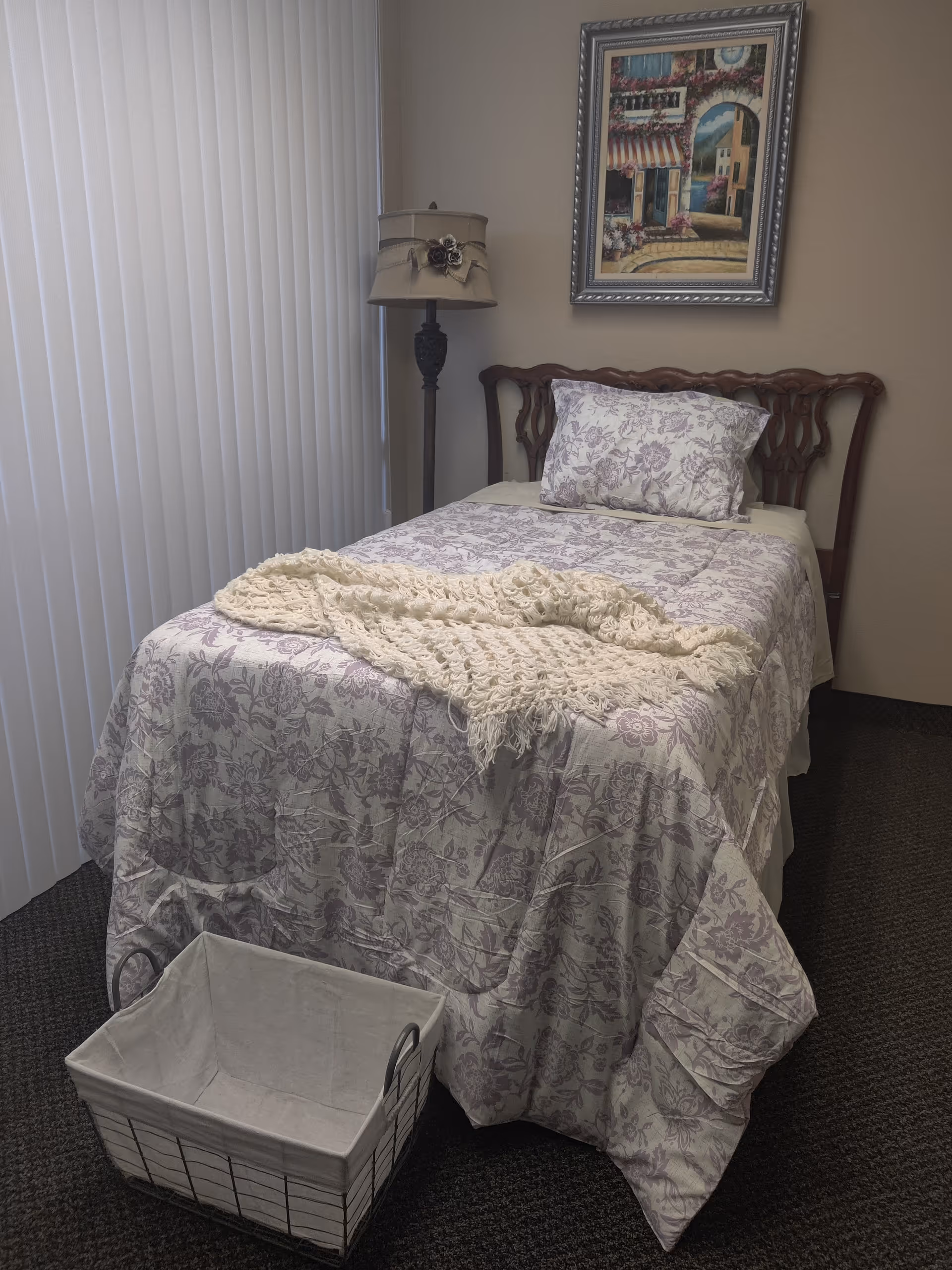 A neatly made bedroom with floral bedspread, lamp, framed artwork, and a white basket.
