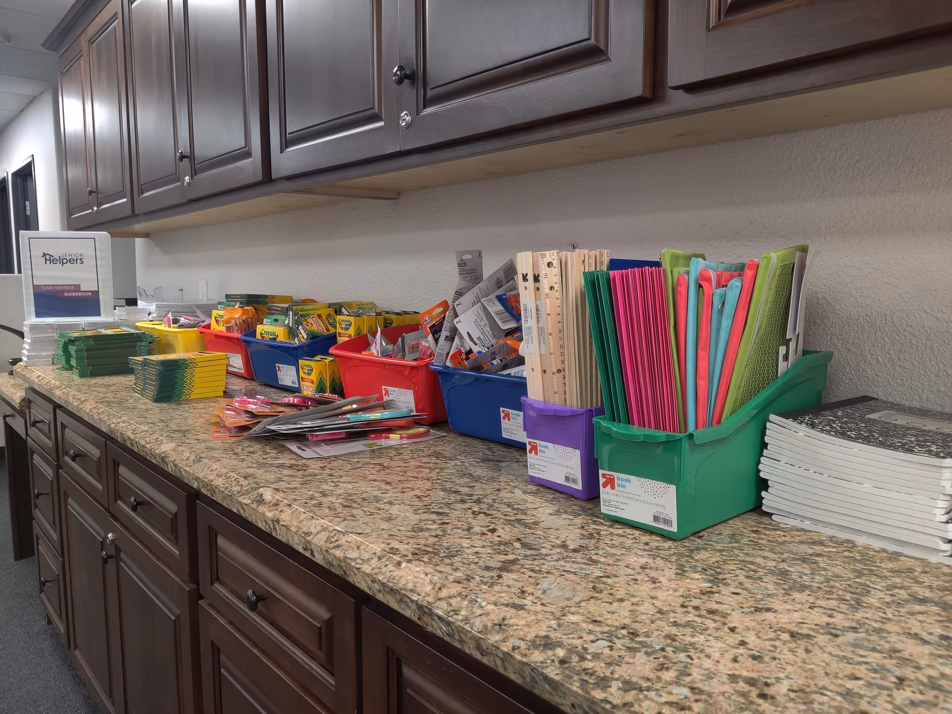 Granite countertop with colorful bins of school supplies beneath dark cabinets.