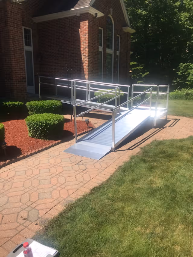 Metal wheelchair ramp leading to brick facility entrance with manicured shrubs.