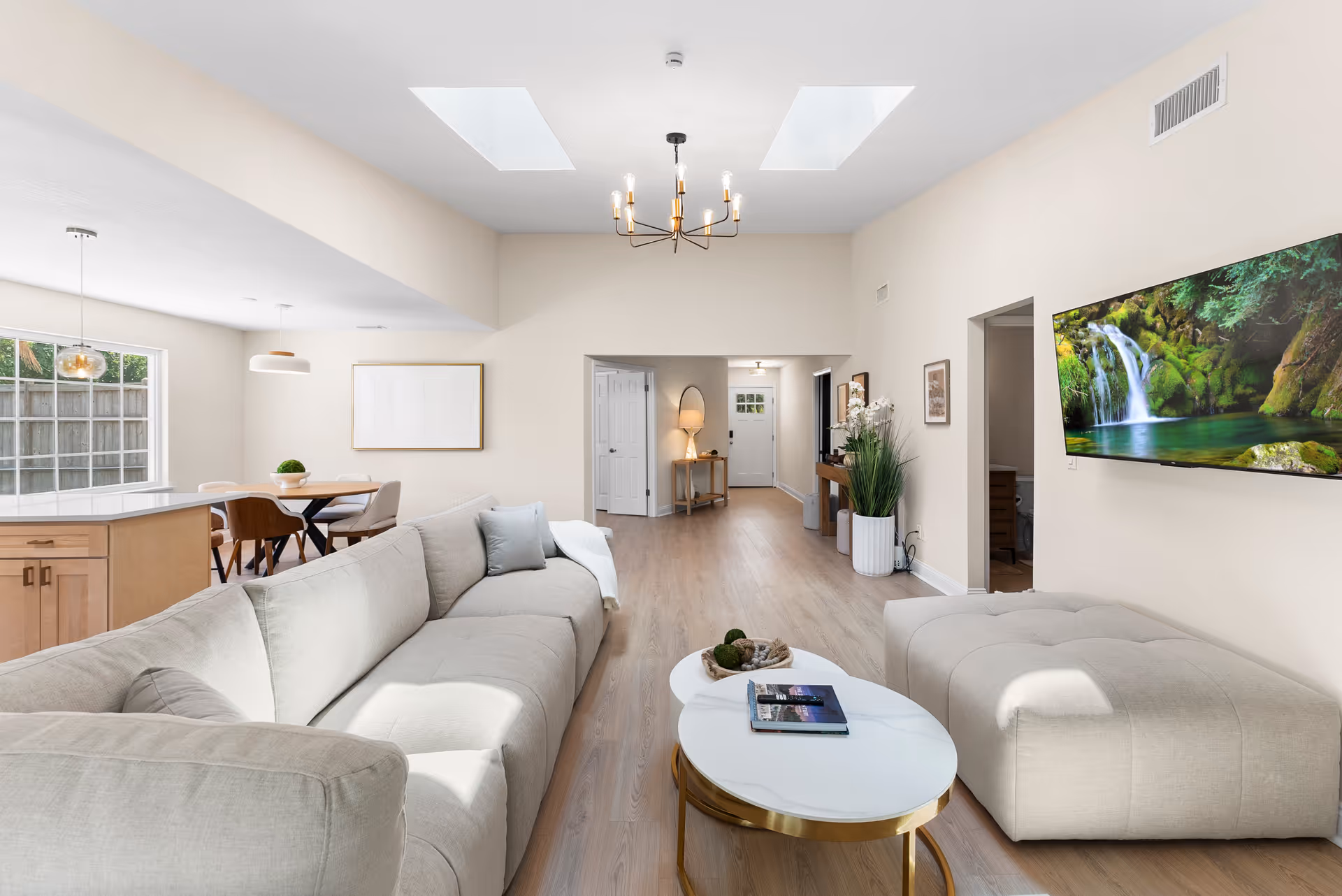 Bright open living room with beige sectional sofa, coffee table, TV, skylights, and dining area.
