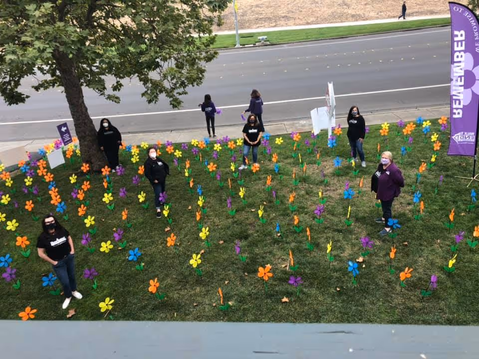 People wearing masks stand on a grassy lawn among colorful pinwheel flowers near a road.