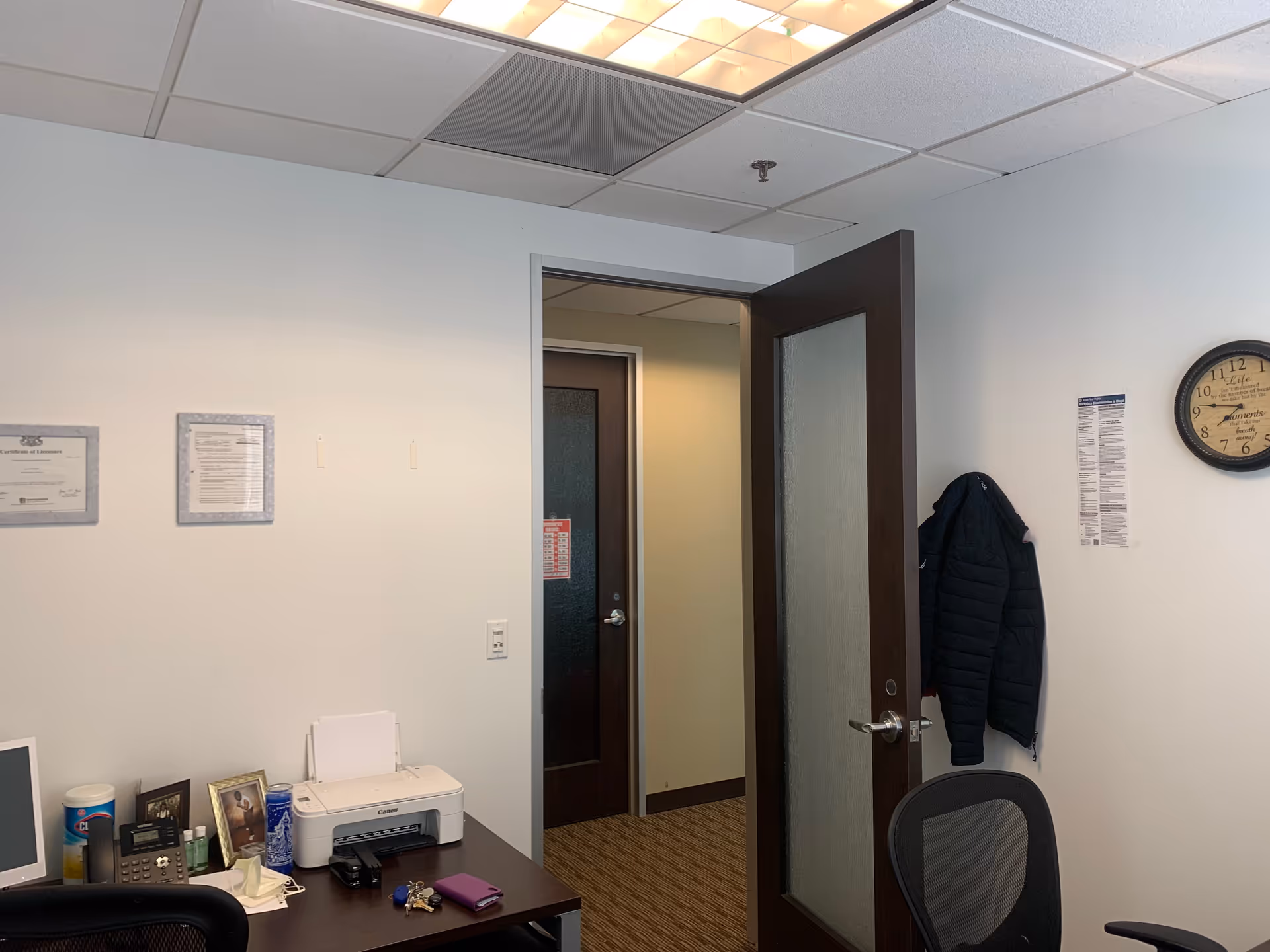 Office corner with desk, printer, jacket on the wall, and an open doorway to hallway.