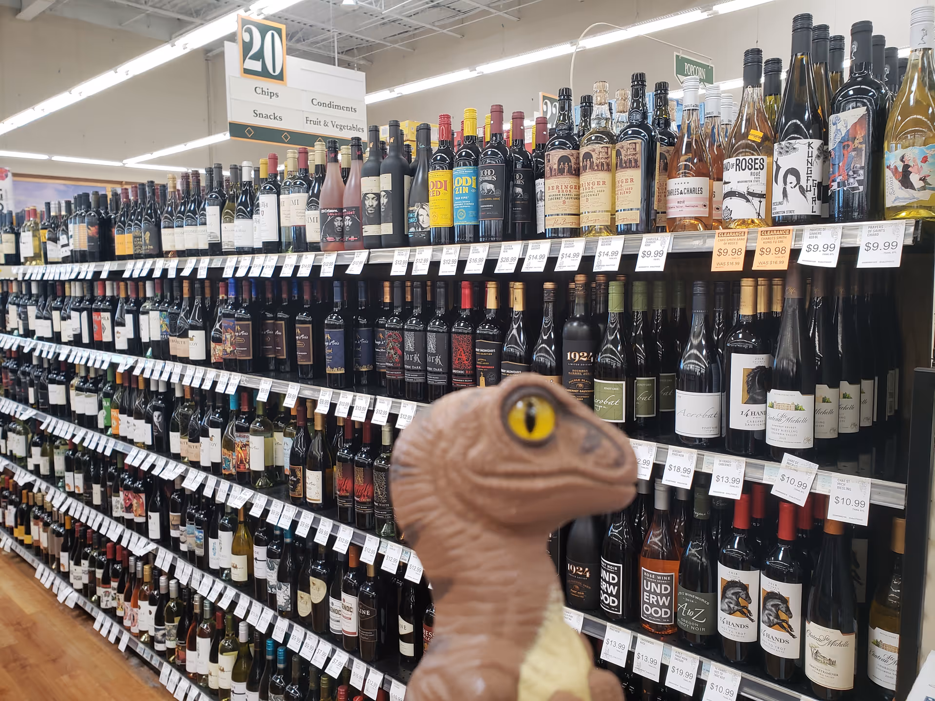 Dinosaur toy in foreground with long shelves of wine bottles in a store aisle.
