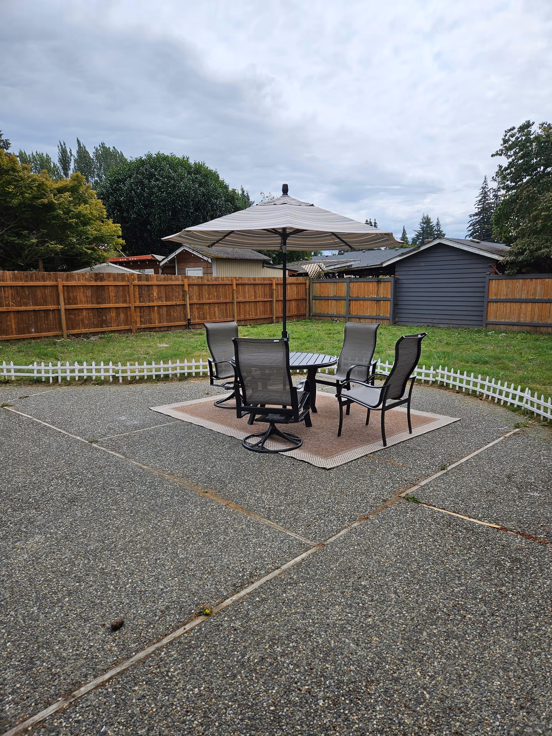 Outdoor patio with a table, chairs, and umbrella on a concrete surface behind a wooden fence.