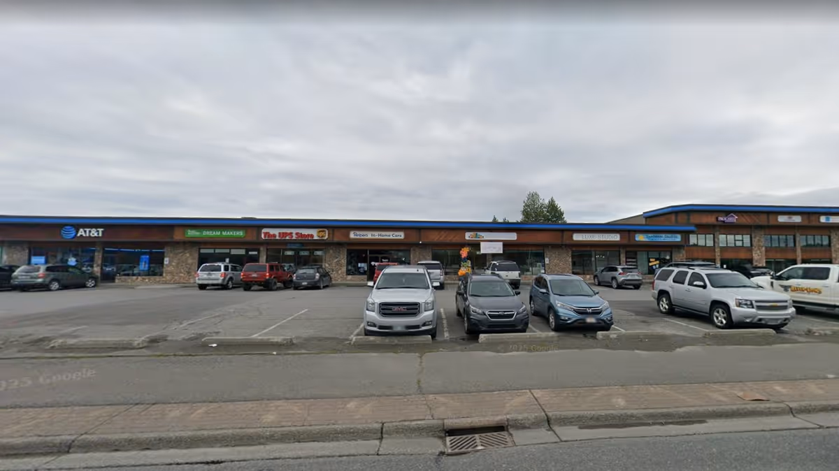 Row of storefronts in a strip mall with parked cars in front.