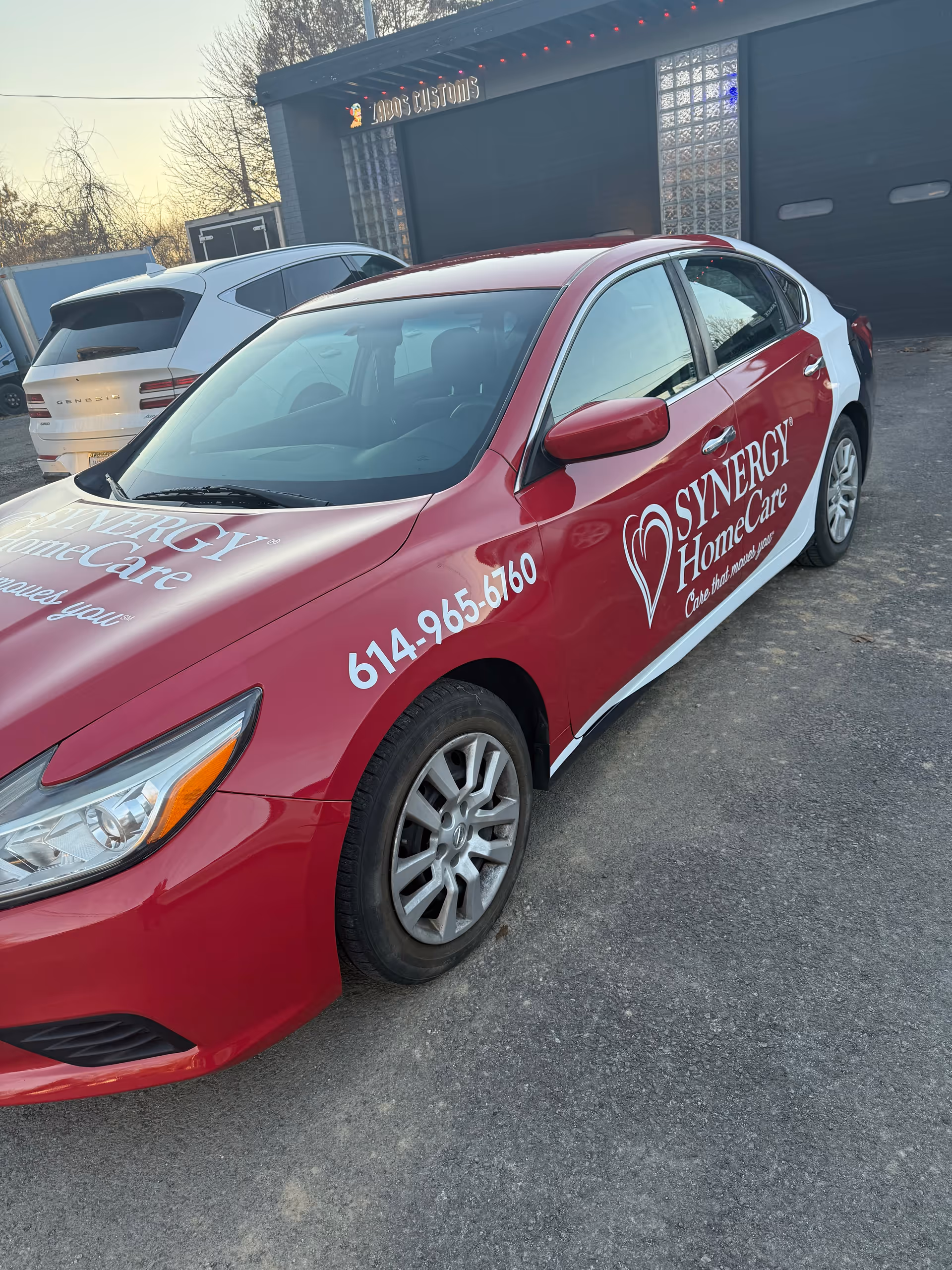 A red Synergy HomeCare car with logo parked in a lot outside a dark building.